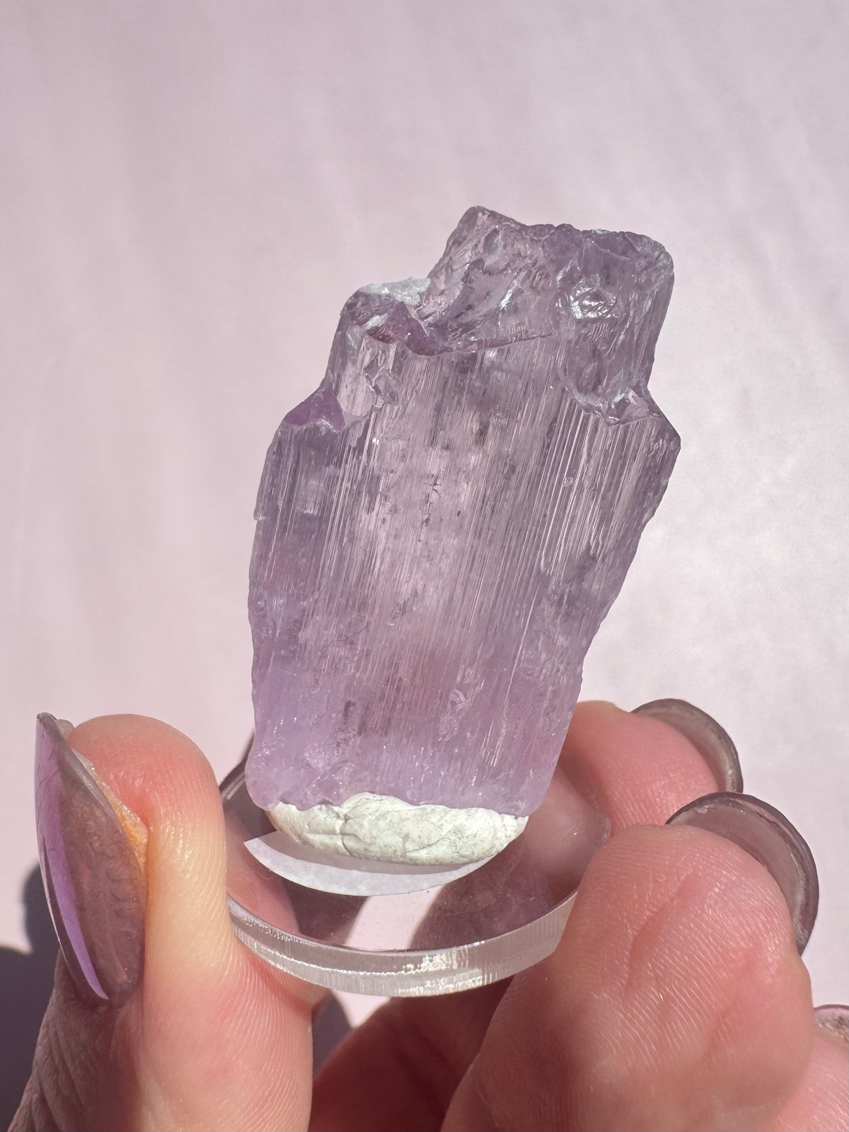A hand displays Lavender Kunzite (Spodumene) - Item A, a pale purple rough crystal upright on a clear circular base, set against a light pink background.