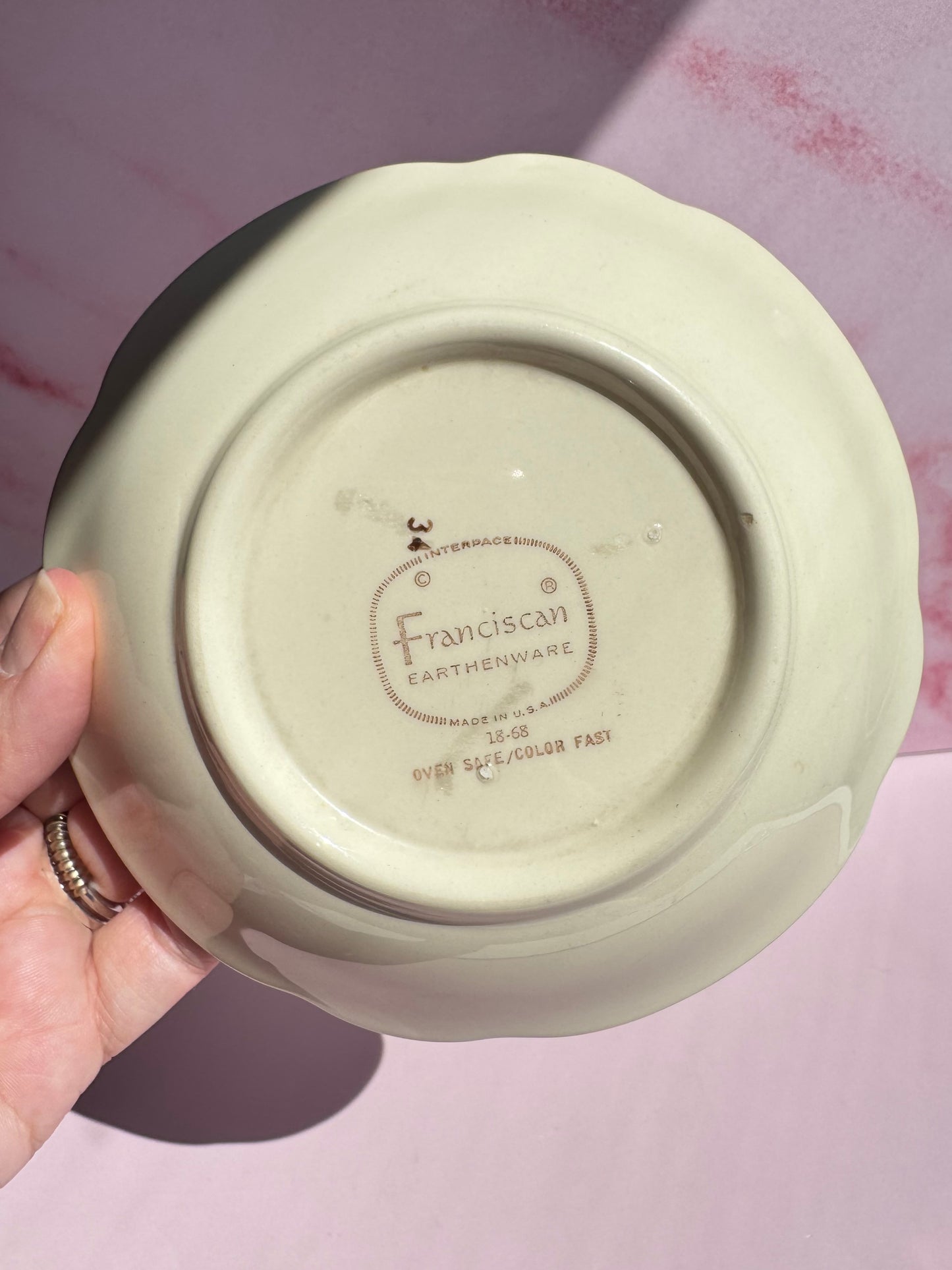 A hand holds a cream-colored Desert Rose Berry Bowl upside down, revealing the "Franciscan Earthenware" maker's mark along with details about its oven safety and color fastness on this vintage floral dish.