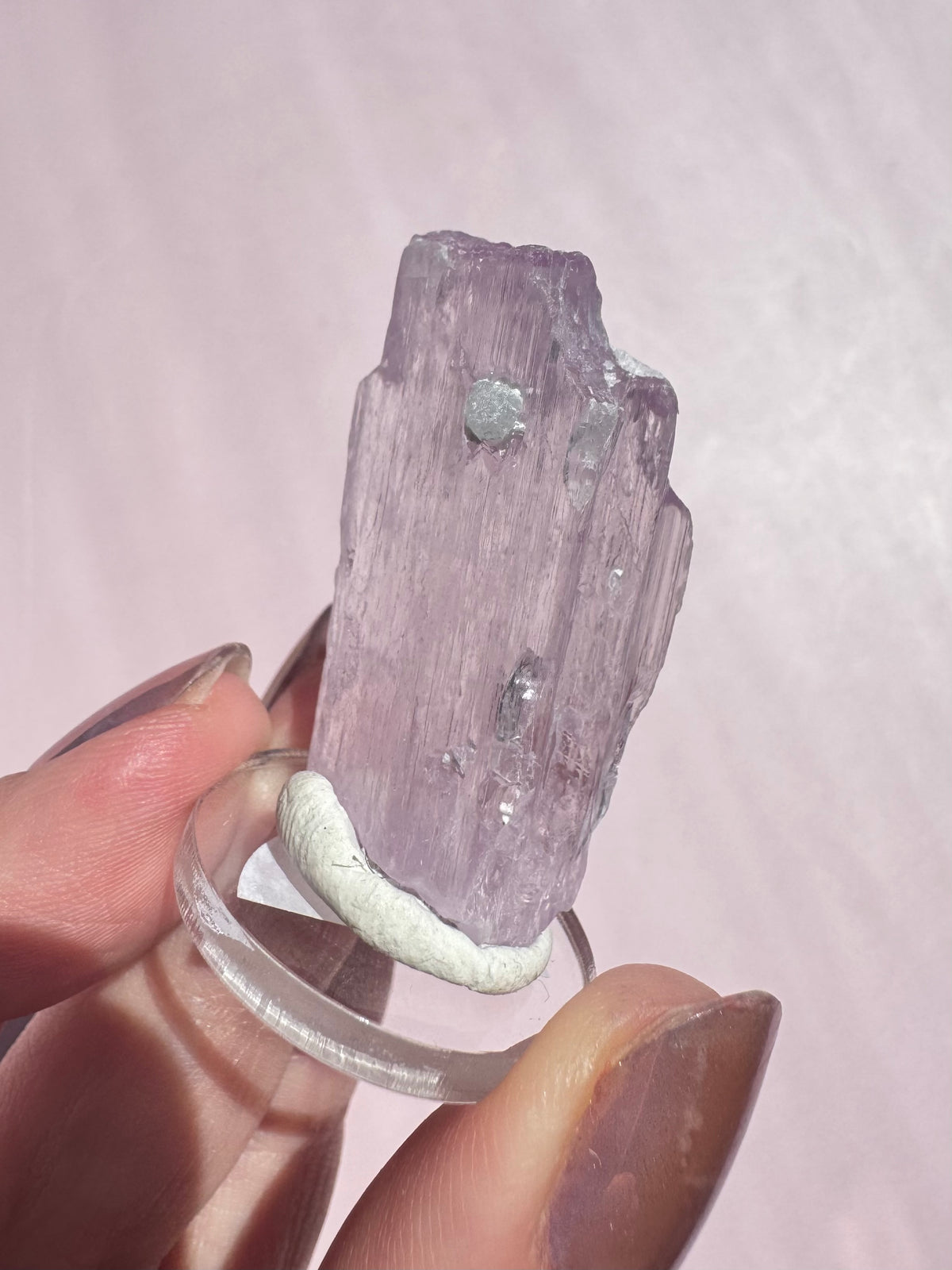A hand holding the Lavender Kunzite (Spodumene) - Item A, a pale purple, translucent raw crystal specimen mounted on a clear display stand with white putty.