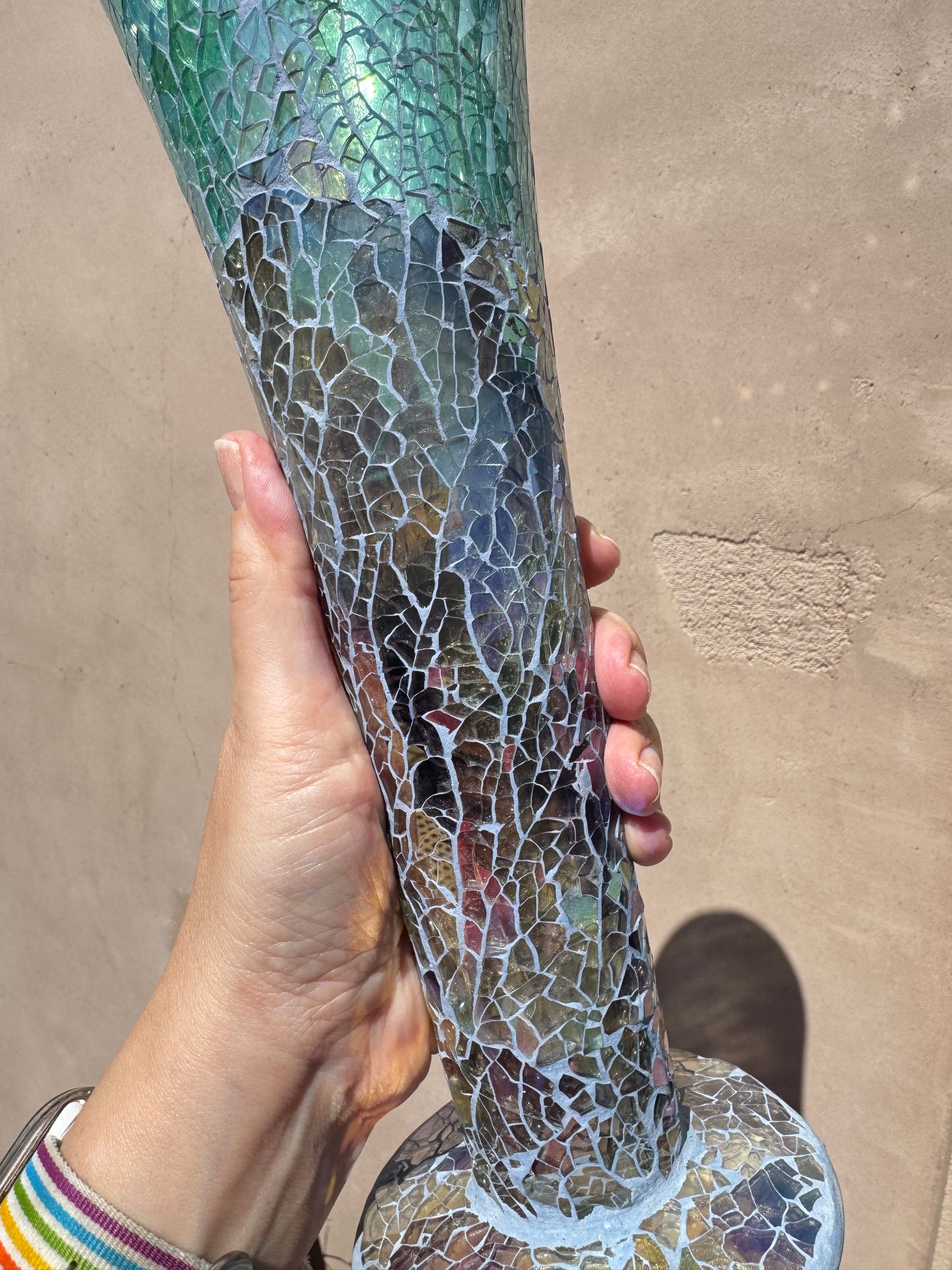 A hand holding the Iridescent Blue Mosaic Crackle Glass Vase, a tall trumpet-shaped piece featuring a crackled mosaic pattern, against a beige wall.