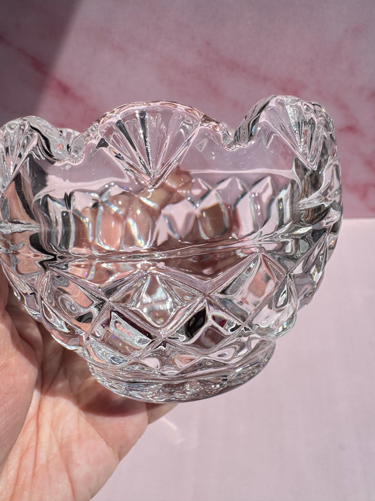 A hand holds the Floral Crystal Bowl—a clear, cut glass piece with a scalloped rim, geometric patterns, and a delicate frosted leaf accent—against a pink background.