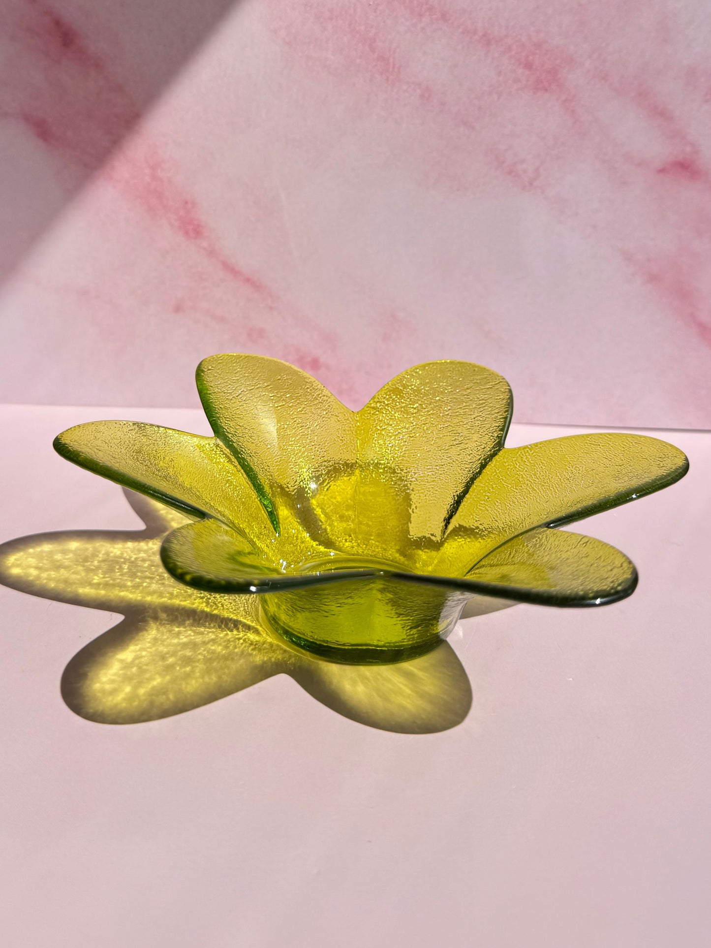 A Green Flower Glass Bowl (Imperfect), with its translucent petals, rests on a pink surface and casts a delicate flower-shaped shadow.