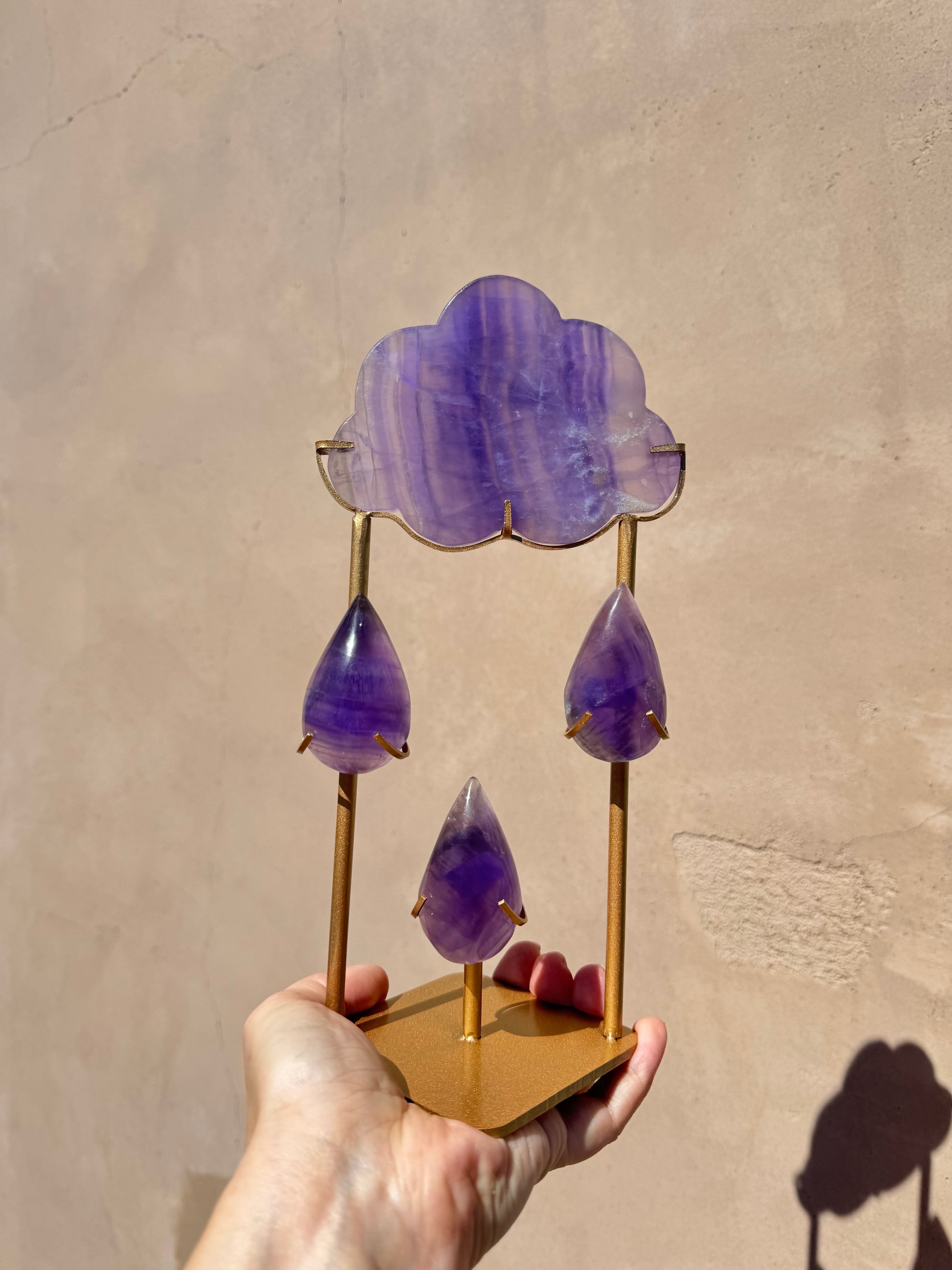 A hand holds the Rain Cloud - Purple Fluorite B, a decorative object featuring a cloud carving made of purple fluorite crystal on top with three purple teardrop shapes hanging below, all mounted on a gold stand against a beige wall.