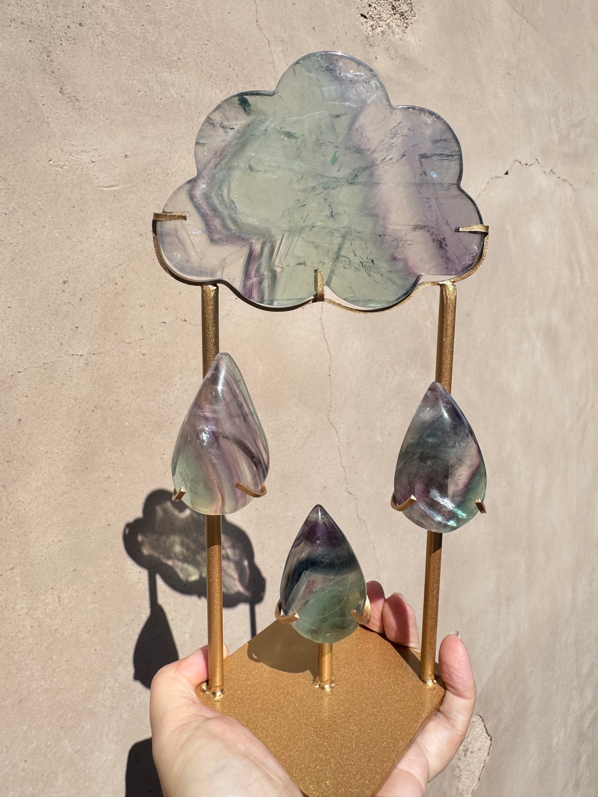 A hand holds the Rain Cloud - Rainbow Fluorite—a gold metal stand featuring a cloud and three raindrops carved from translucent, multicolored fluorite—in sunlight, casting a shadow on a beige wall.