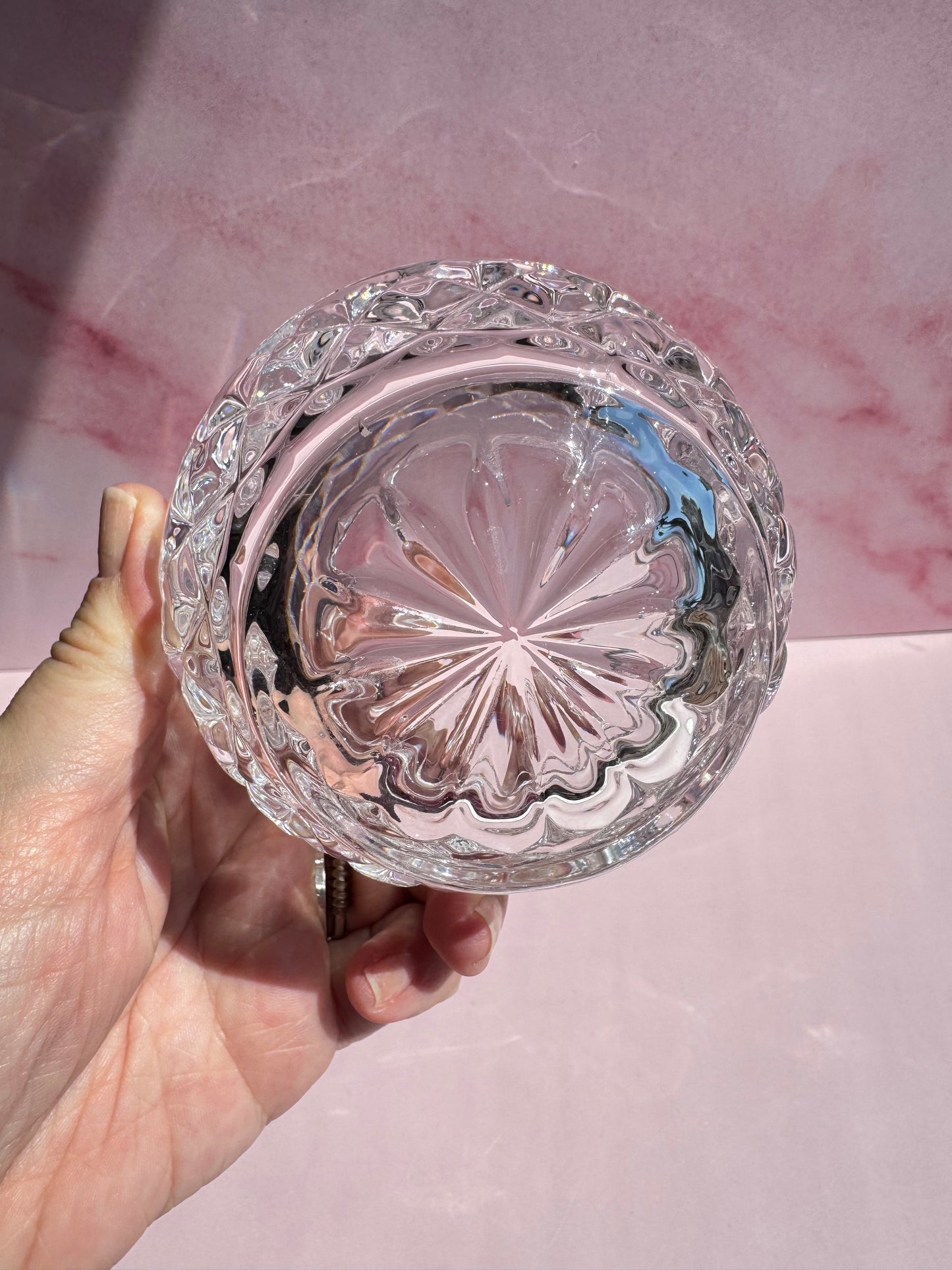 A hand holds the Floral Crystal Bowl, a round, clear glass dish featuring a decorative starburst pattern on the bottom, set against a pink marbled background and evoking the elegance of fine crystal.