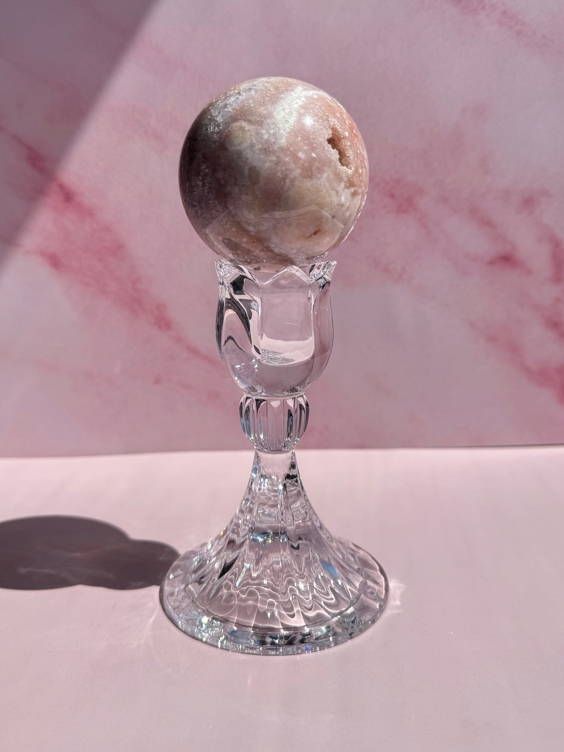 A round, marbled stone sphere is displayed on a clear, ornate Tulip Candle Holder against a pink, marbled background with soft shadows.
