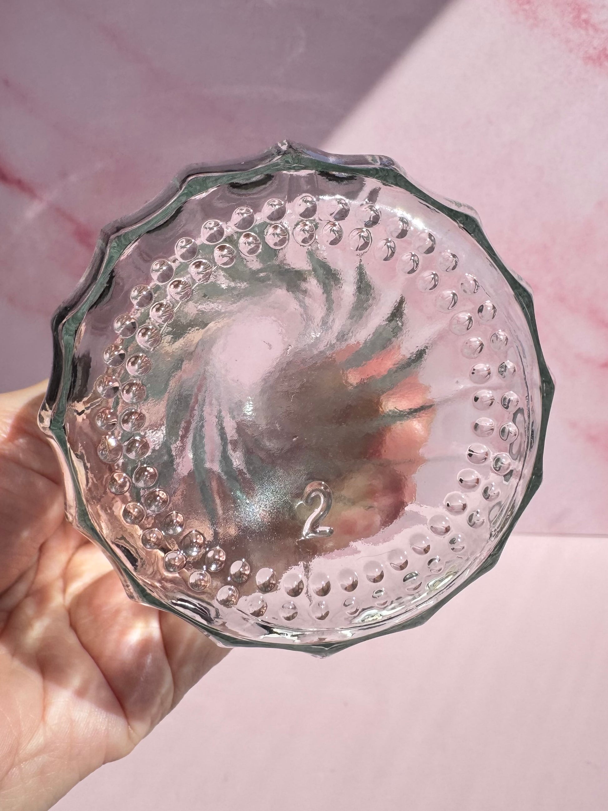 A hand holds the Glass Vase with Spiral Pattern, featuring a clear glass design with a textured, swirled bottom and the number 2 embossed at its center, against a pink background.