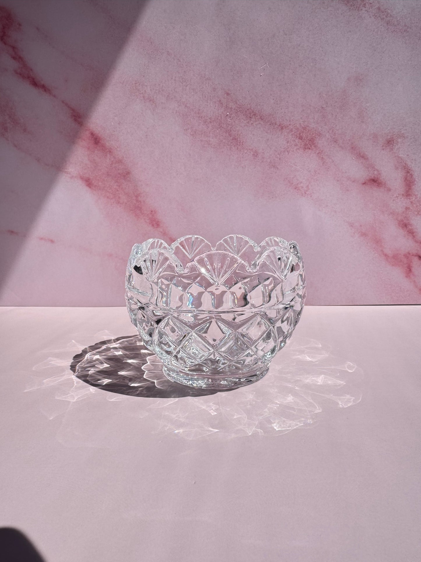 The Floral Crystal Bowl, featuring a delicate flower pattern and crafted from clear glass, sits on a pink surface against a pink marbled background, casting an intricate shadow in bright light.