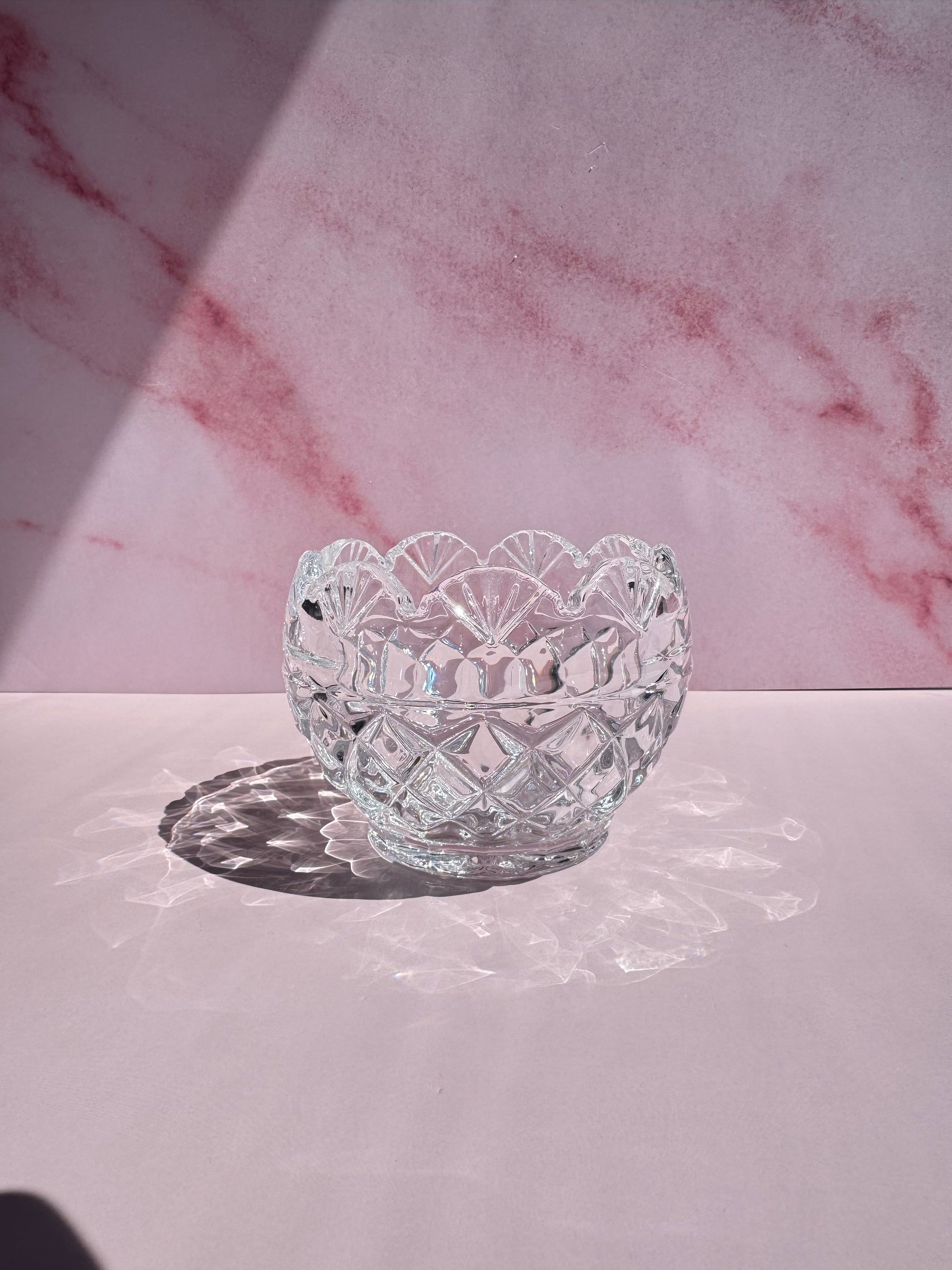 The Floral Crystal Bowl, featuring a delicate flower pattern and crafted from clear glass, sits on a pink surface against a pink marbled background, casting an intricate shadow in bright light.