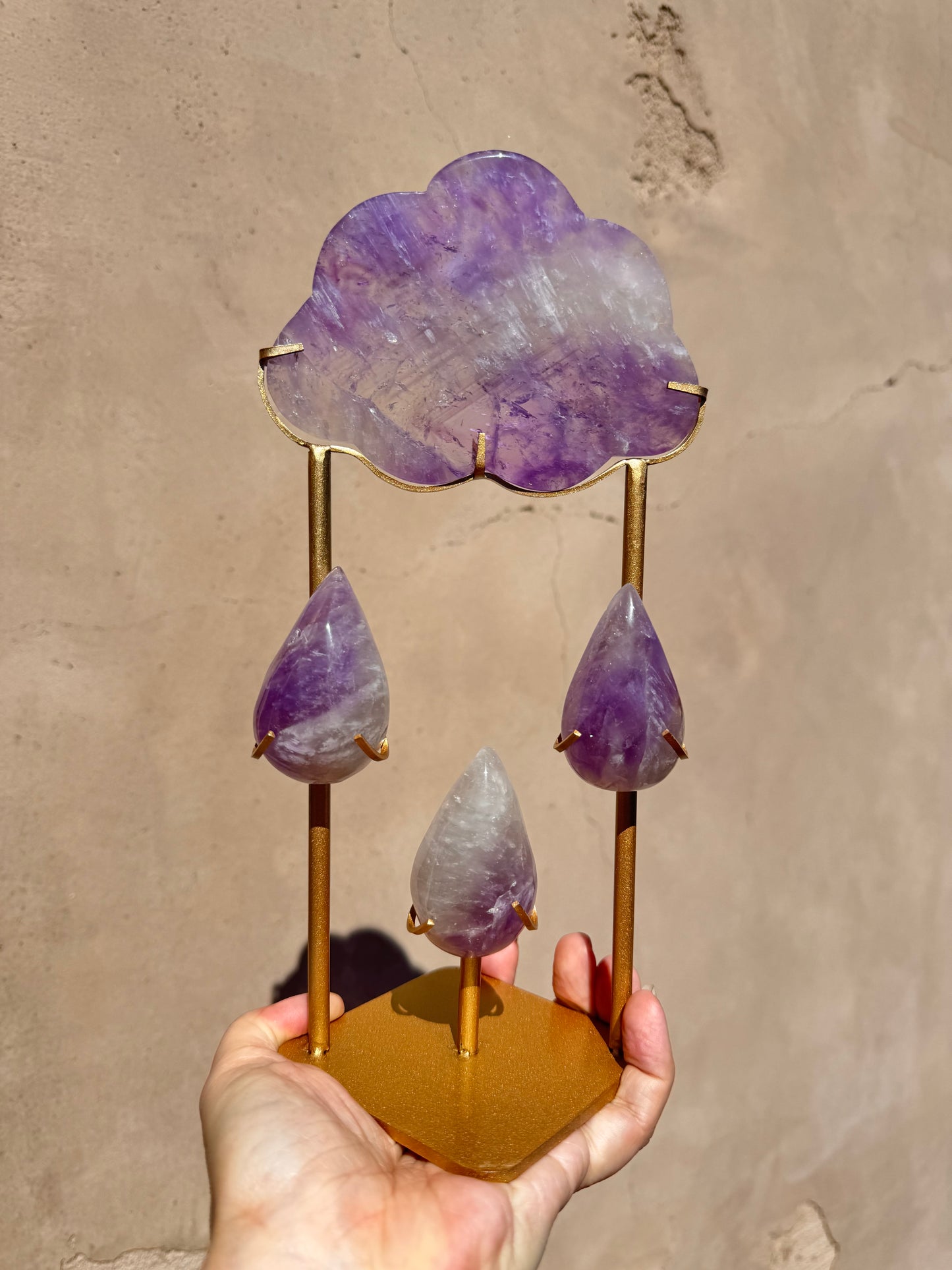 A hand holds the "Rain Cloud - Purple Fluorite A," a decorative piece showcasing cloud and raindrop-shaped purple fluorite carvings elegantly mounted on gold metal stands, displayed against a beige wall and radiating an aura of crystal healing.