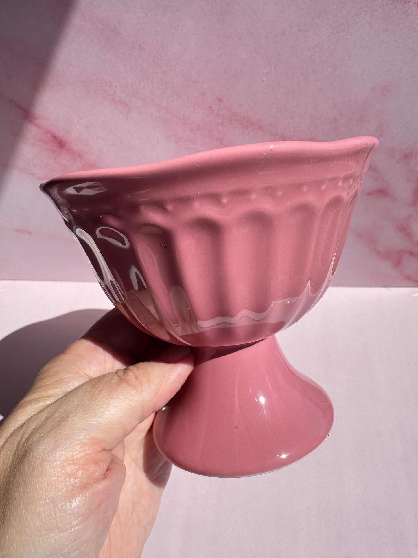 A hand holds the Pink Ceramic Raised Bowl, featuring a glossy finish and delicate scallop rim, against a pink marble-patterned background.