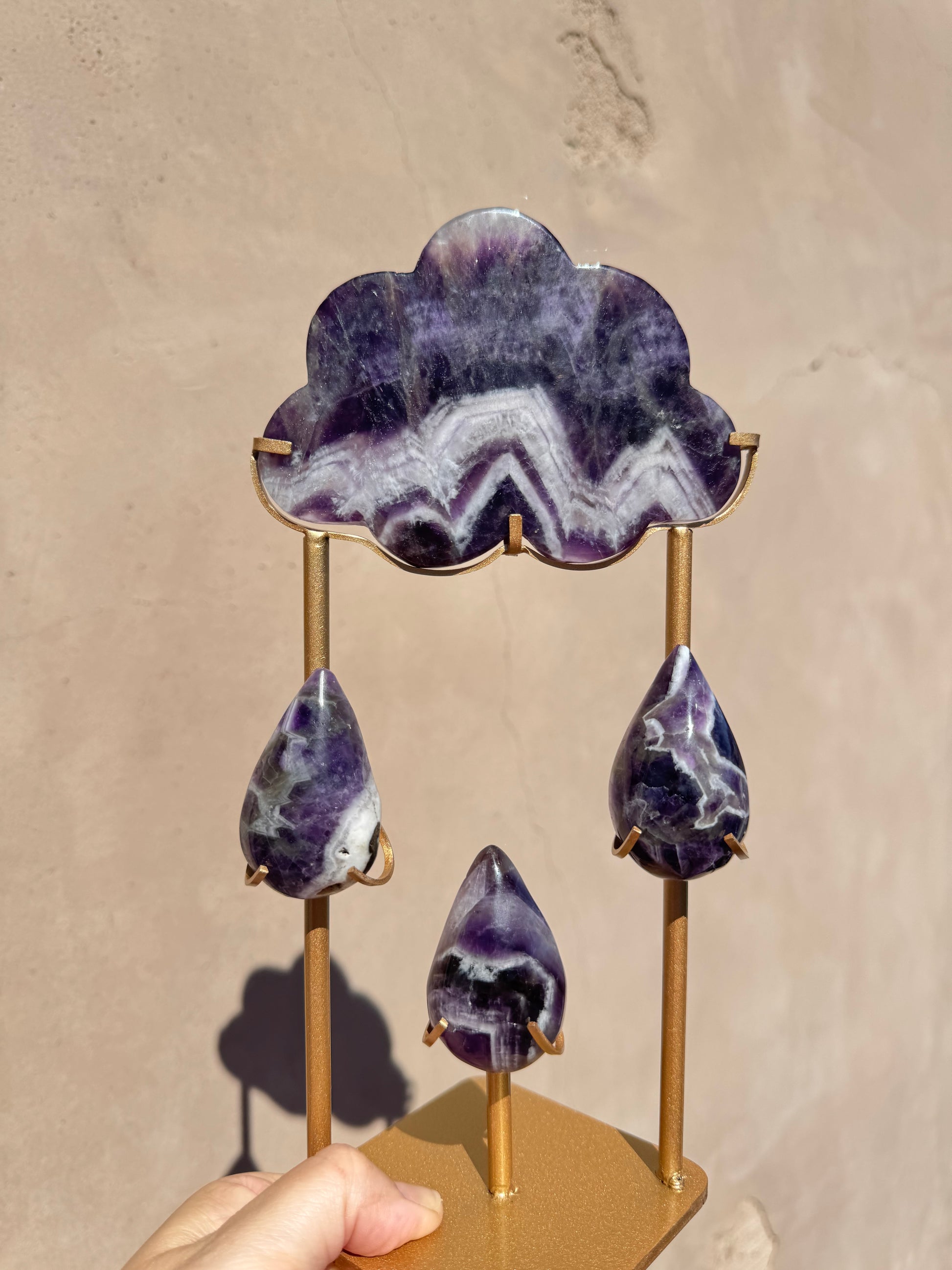 A hand holds the "Rain Cloud - Chevron Amethyst," a metal stand showcasing four purple and white banded chevron amethyst stone pieces shaped like a cloud and three raindrops, set against a beige background.
