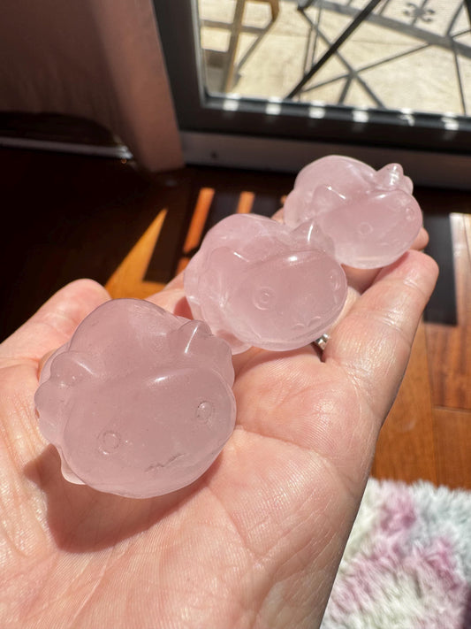 A hand holds three small Rose Quartz Axolotl 2" Carvings—pink crystal axolotl figures—glistening in sunlight by a window.