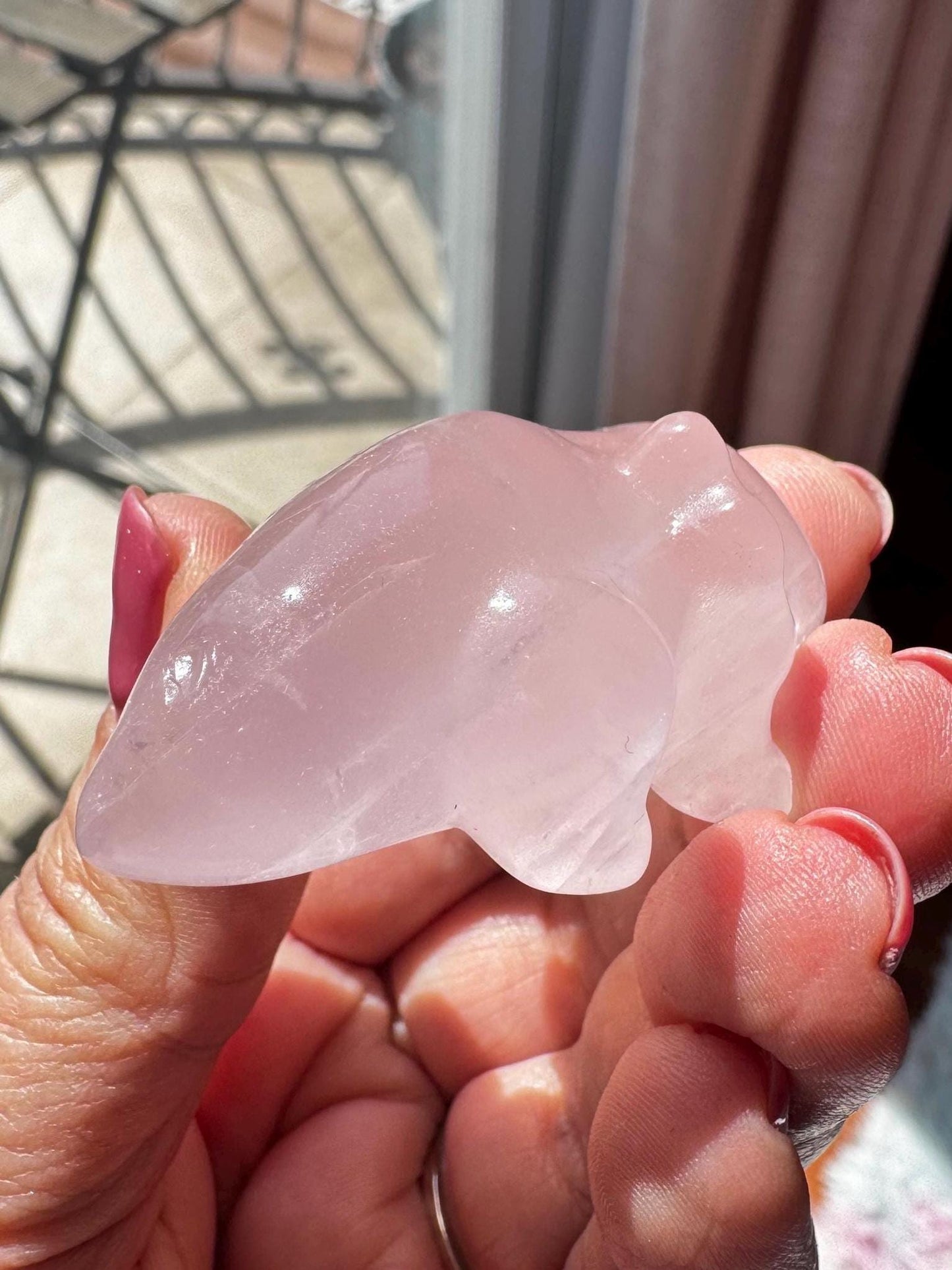 A hand holds a smooth, 2" rose quartz axolotl carving, its pink crystal shimmering in the sunlight—an ideal gift for healing crystal enthusiasts.
