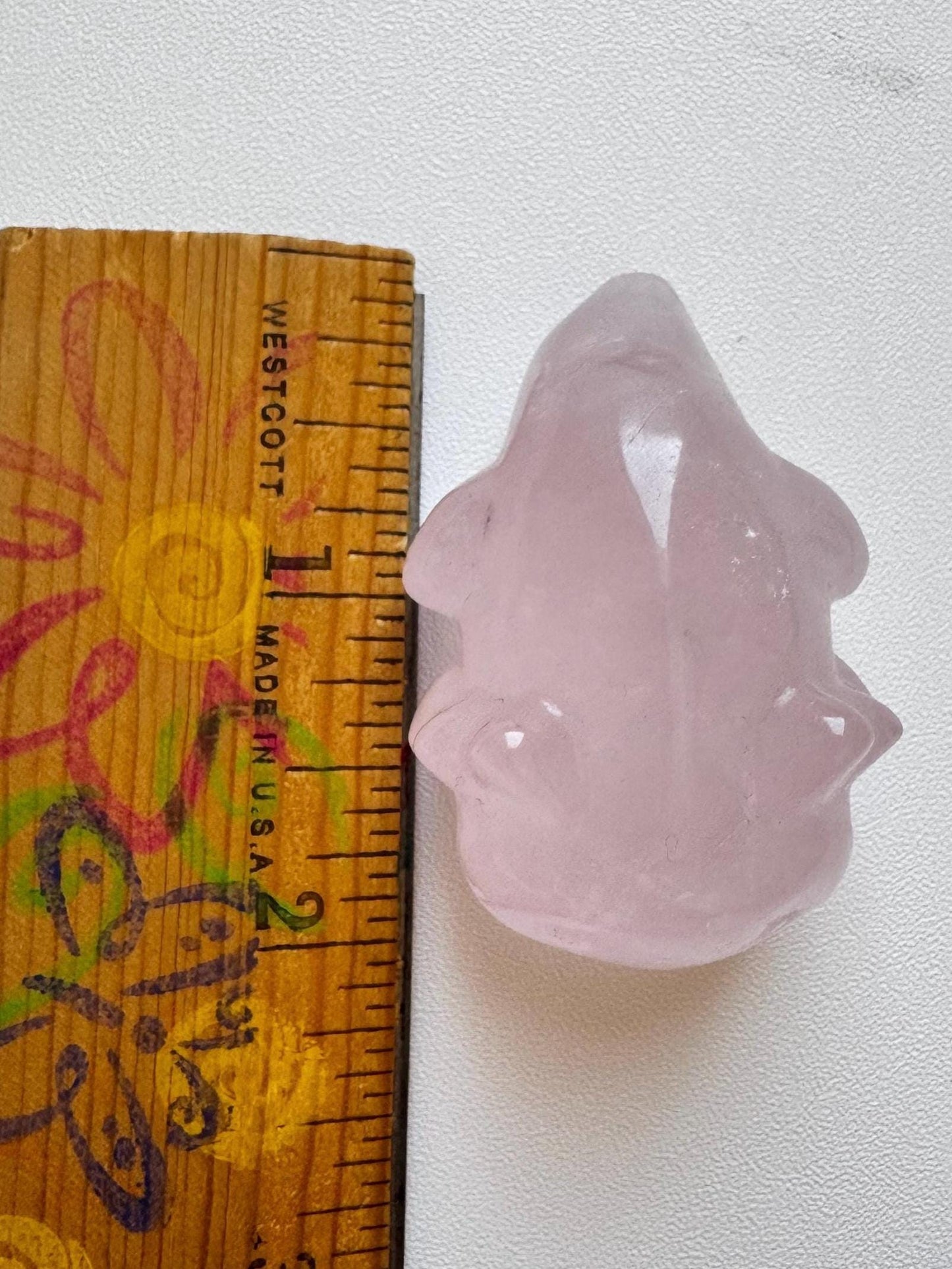 A 2" Rose Quartz Axolotl carving—a pink crystal figure—sits beside a wooden ruler, making a charming gift for fans of healing crystals.