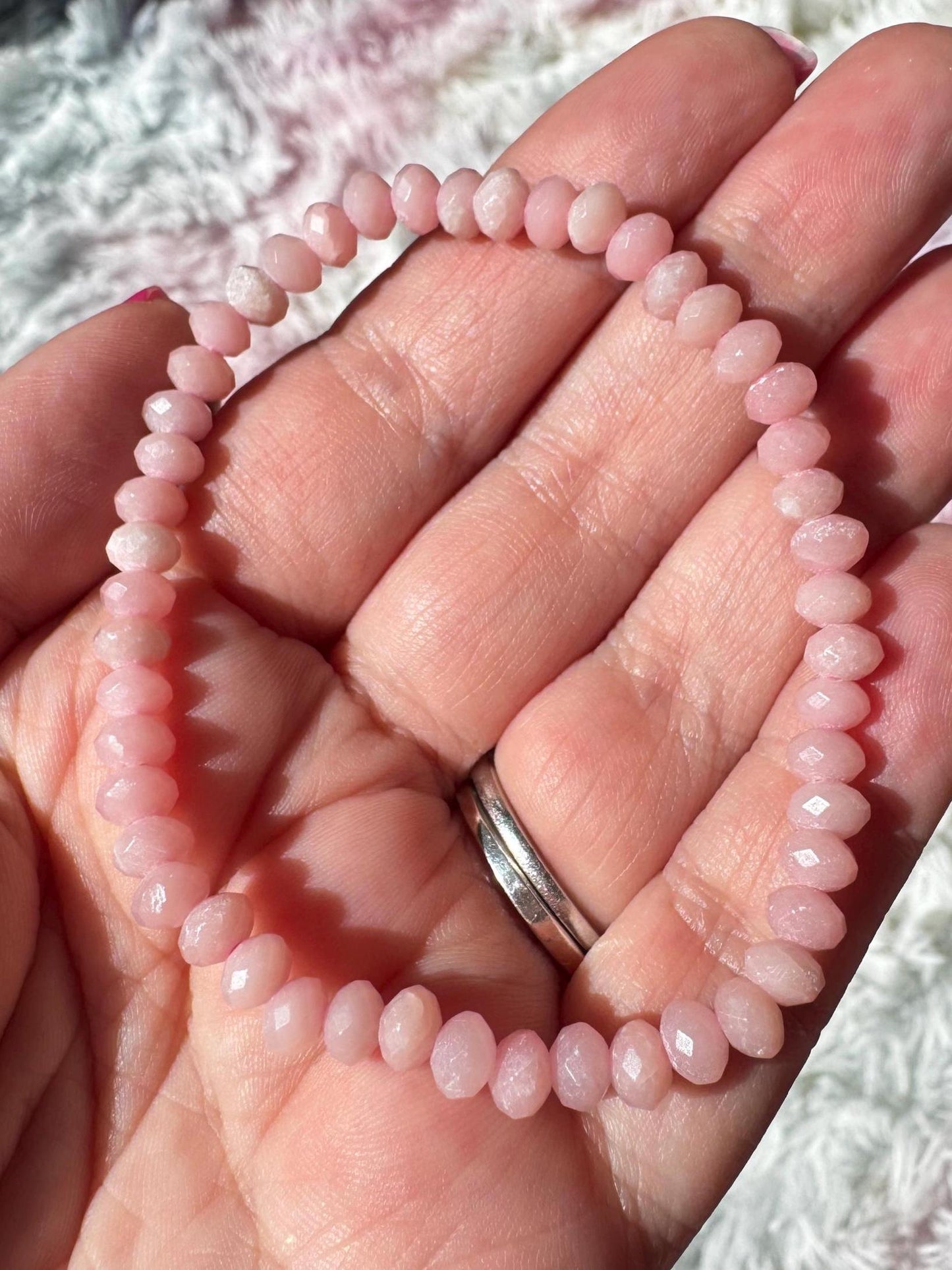 A hand displays the Pink Opal Bracelet, a 4mm stretchy piece with faceted pale pink beads. Two silver rings adorn the ring finger, while a soft background highlights this calming crystal jewelry.