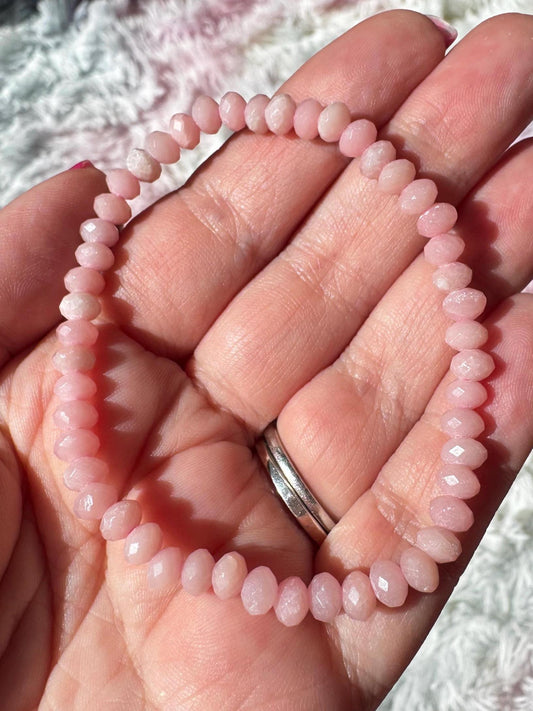 A hand displays the Pink Opal Bracelet, a 4mm stretchy piece with faceted pale pink beads. Two silver rings adorn the ring finger, while a soft background highlights this calming crystal jewelry.