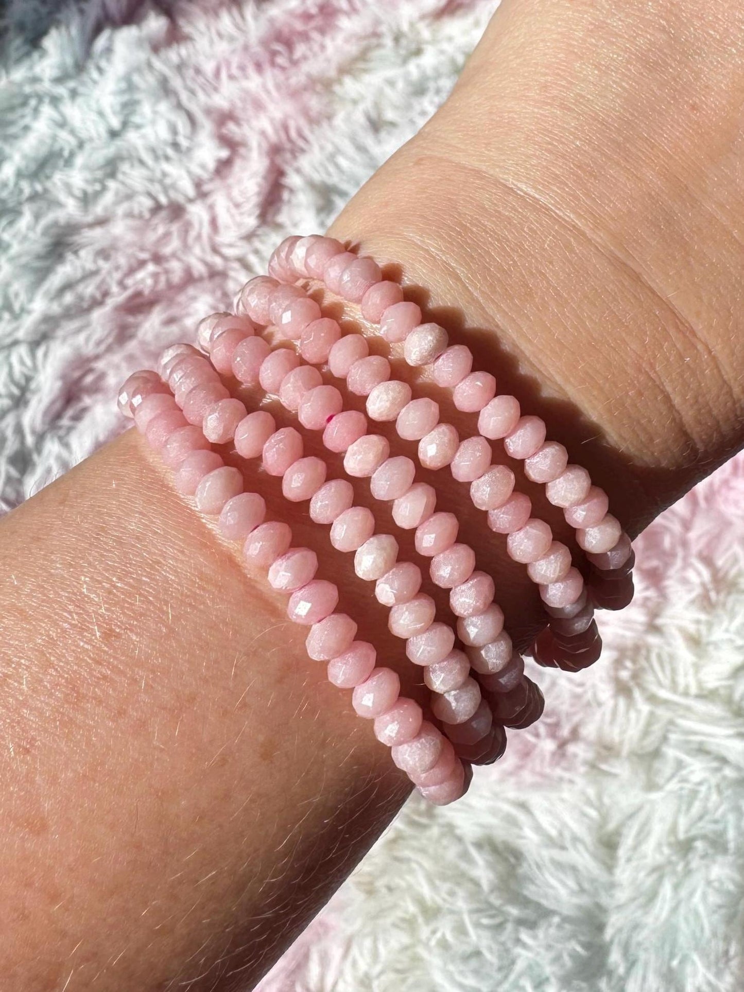 A wrist is shown wearing six stacked, round, faceted light pink beaded bracelets—highlighting the Pink Opal Bracelet, a 4mm stretchy bracelet—against a soft, textured white background.