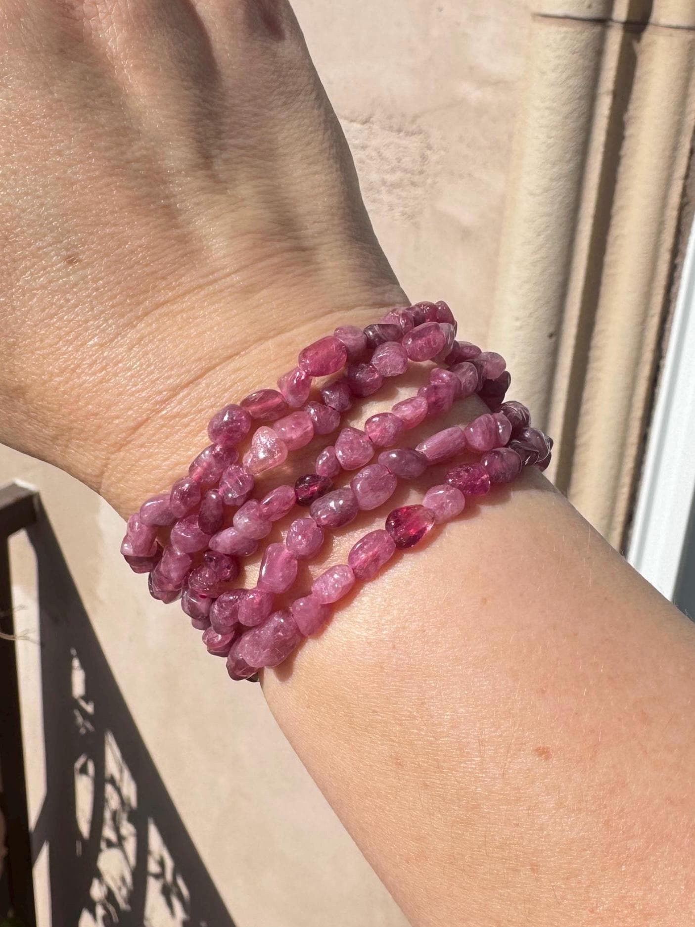 A hand adorned with several pinkish-purple beaded bracelets, including the Pink Tourmaline Chip Bracelet (7" Stretchy, Dark Pink Crystal), is displayed outdoors, highlighting its Heart Chakra and crystal healing benefits.