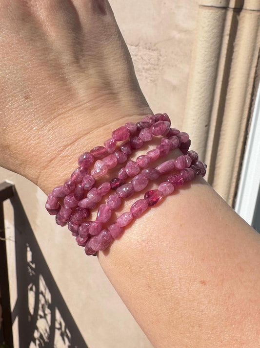 A hand adorned with several pinkish-purple beaded bracelets, including the Pink Tourmaline Chip Bracelet (7" Stretchy, Dark Pink Crystal), is displayed outdoors, highlighting its Heart Chakra and crystal healing benefits.