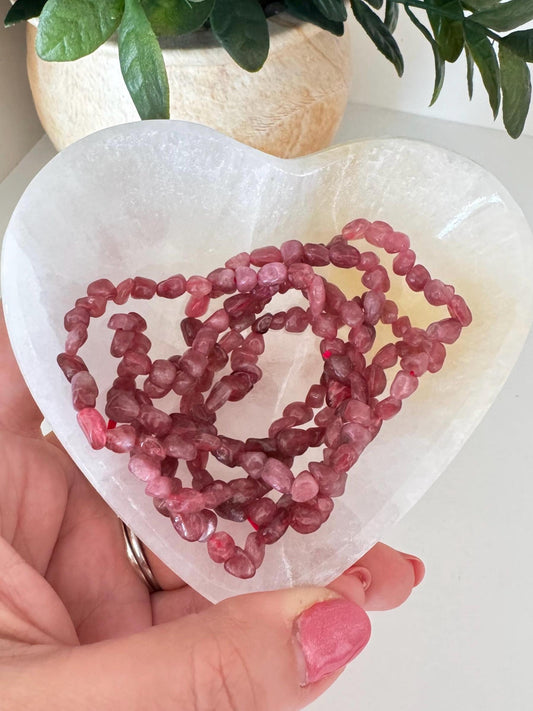 A hand holds a heart-shaped white dish with a Pink Tourmaline Chip Bracelet—7" stretchy, dark pink crystal beads—perfect for crystal healing, with a potted plant in the background.