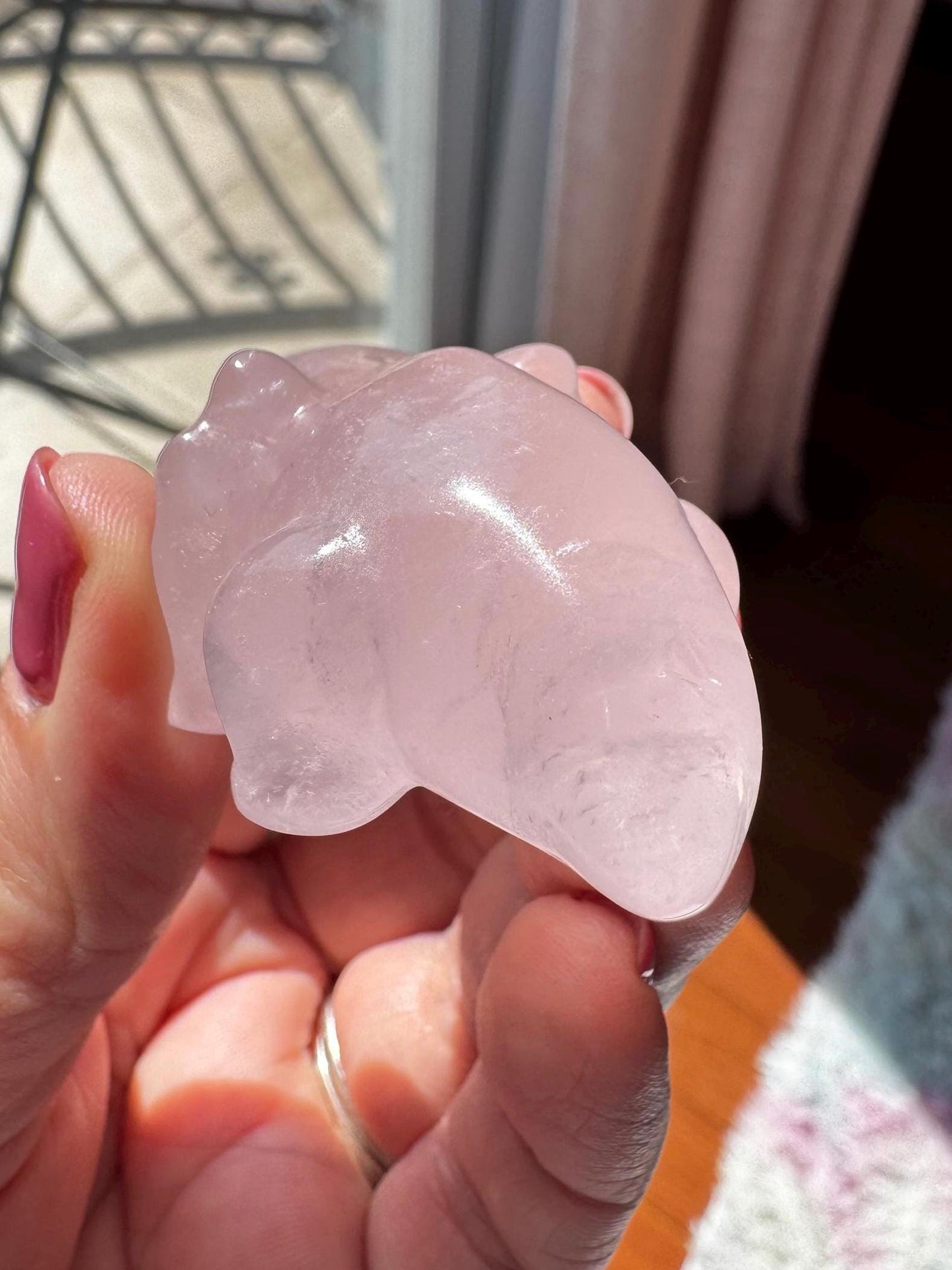 A hand holds a polished, translucent Rose Quartz Axolotl 2" Carving, with sunlight shining on it indoors—a charming pink crystal gift for healing crystal enthusiasts.