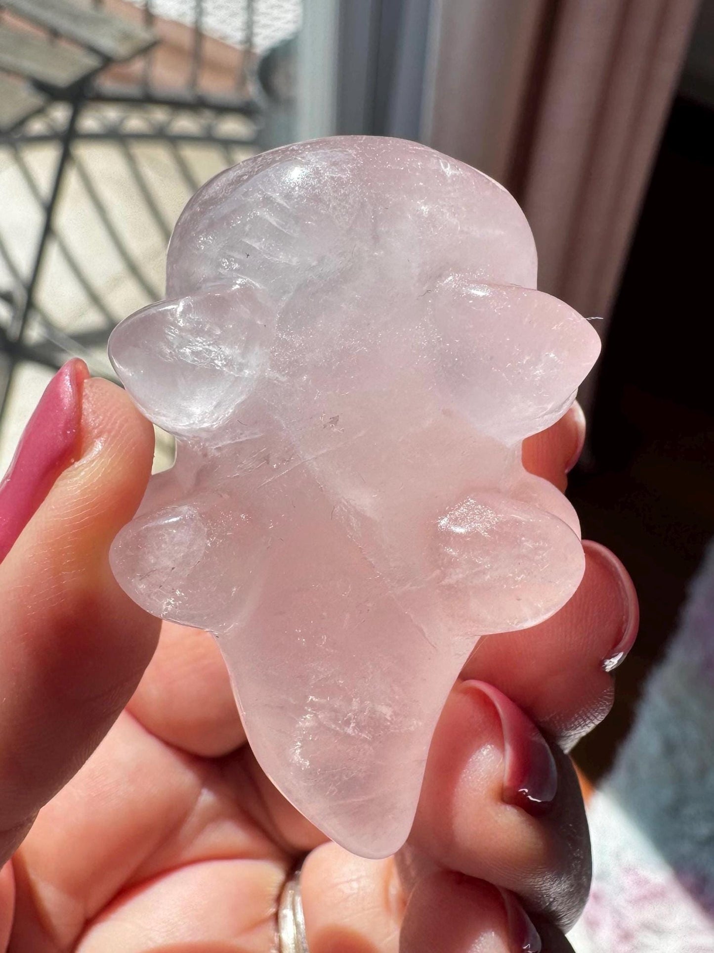 A hand displays a polished Rose Quartz Axolotl 2" Carving—an abstract pink crystal axolotl—glowing in sunlight near a window; perfect as a Rose Quartz Crystal Gift.