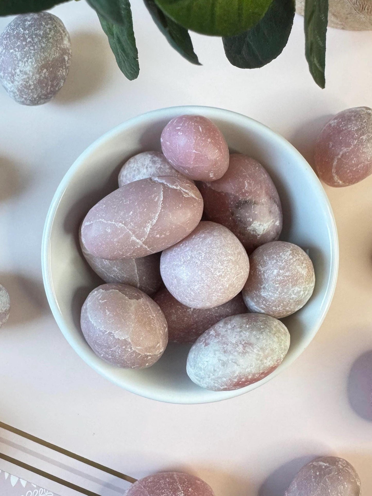 A white bowl with several Pink Opal Tumbles from Peru rests on a light surface, surrounded by more of these soft pink crystal stones—perfect for meditation or as pocket stones, cherished for their gentle beauty and emotional healing properties.