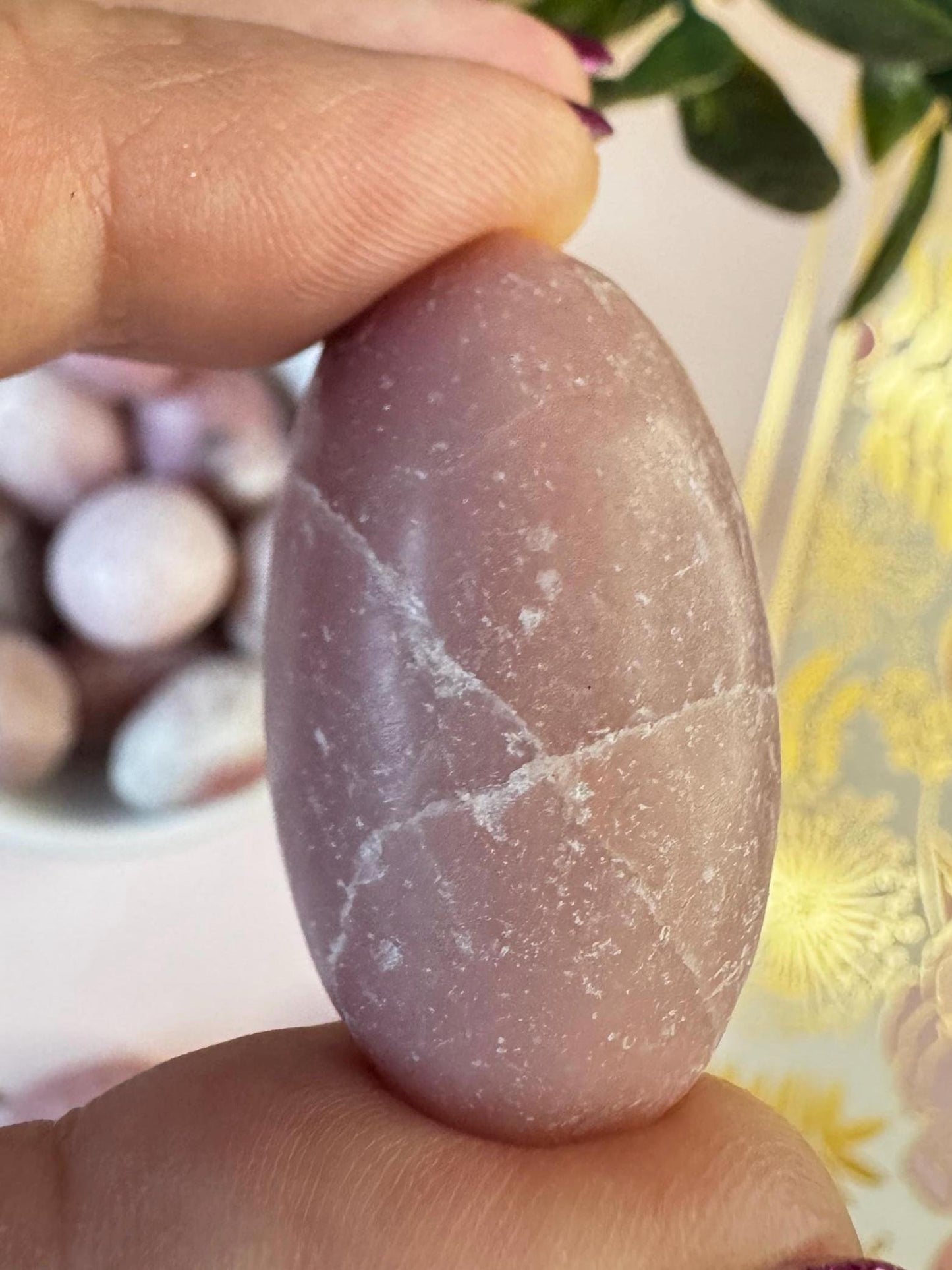 A hand holds a polished Pink Opal Tumble from Peru, showing soft pink hues and white streaks; blurred healing crystals appear in the background. This beautiful crystal is perfect as a pocket stone or for meditation.
