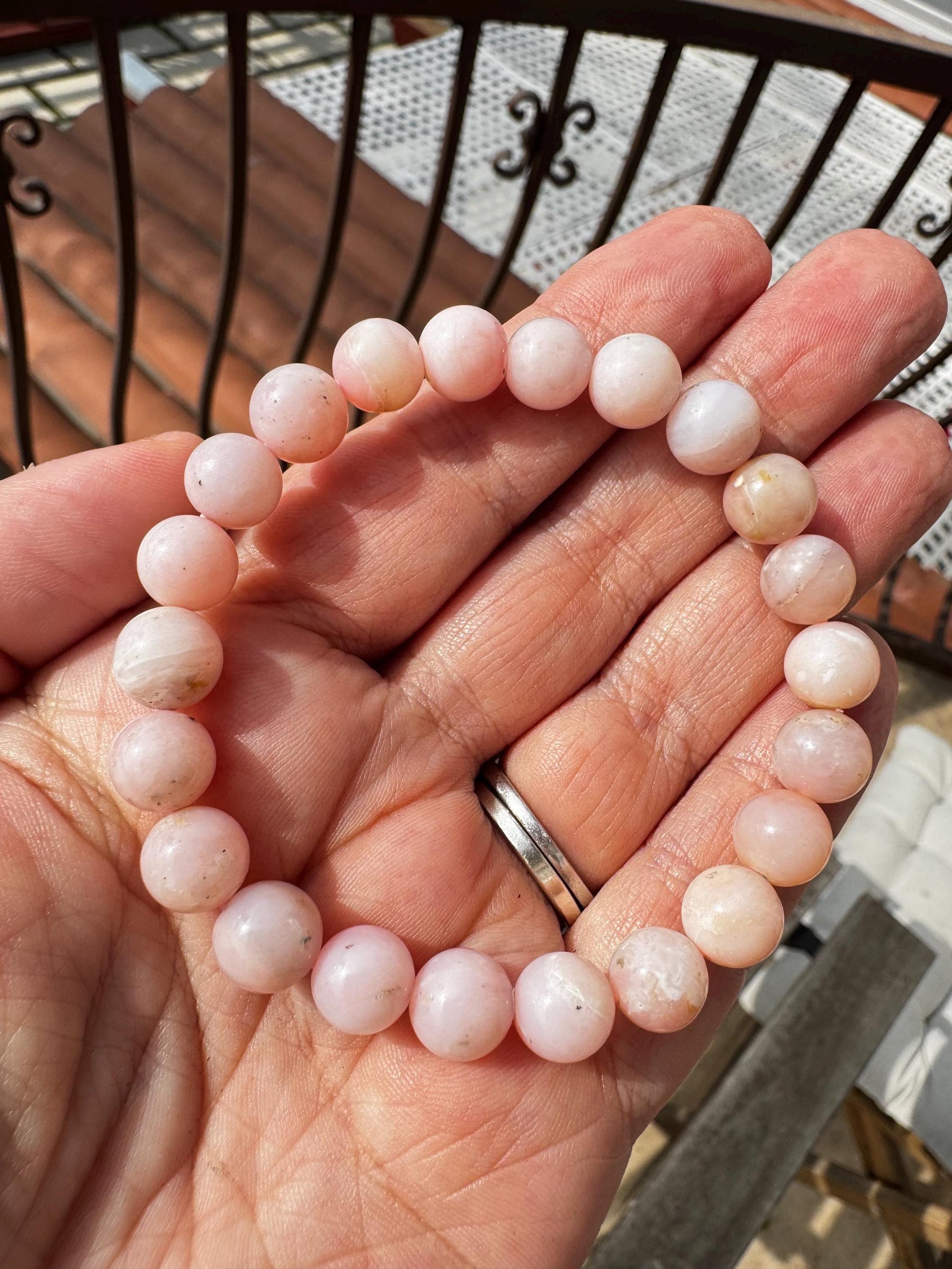 A hand holds a Pink Opalite Bracelet—an 8mm stretchy beaded, man-made crystal bracelet in 6" size—with a metal railing and stairs visible in the background.