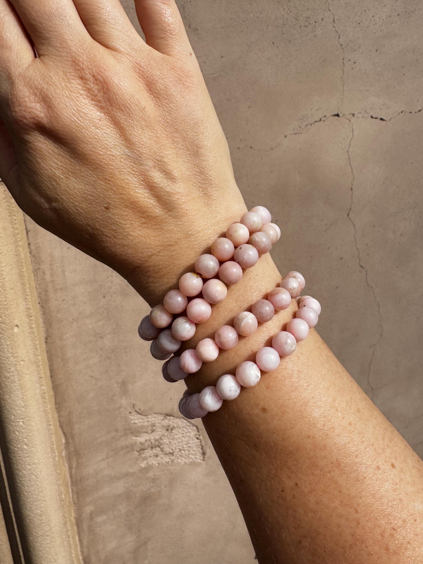 A hand wearing four pink beaded bracelets, including a Pink Opalite Bracelet (8mm stretchy, man-made crystal, 6"), is shown against a beige textured wall with visible cracks.