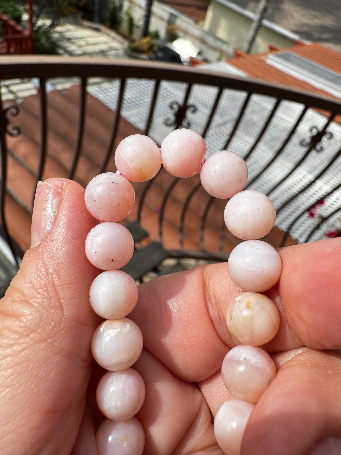A hand holds a Pink Opalite Bracelet (8mm stretchy beads, 6" size, man-made crystal), believed to support emotional balance, with an outdoor balcony and rooftops in the background.