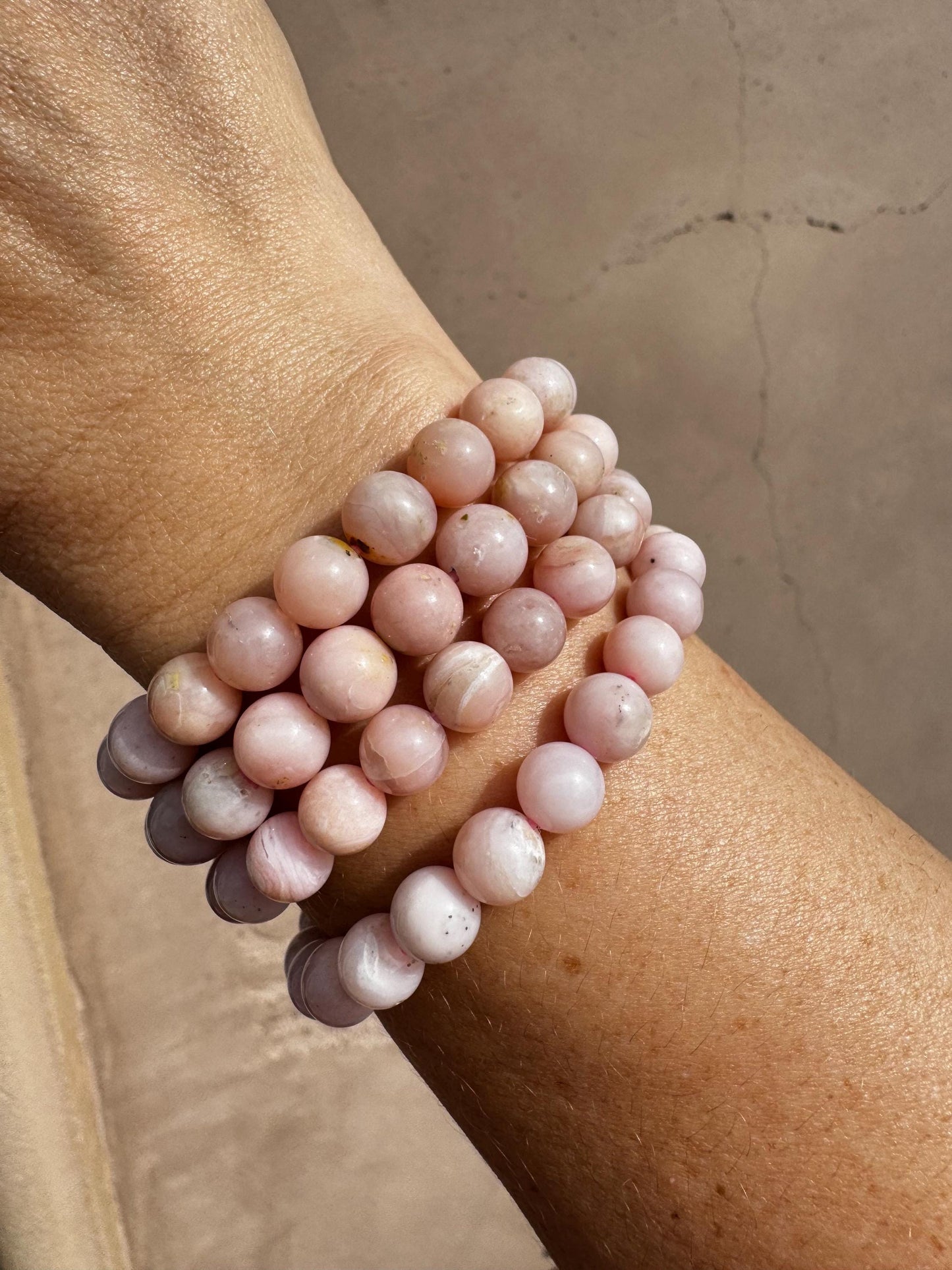 A close-up of a wrist wearing three pink beaded bracelets, including a 6" Pink Opalite Bracelet made with 8mm man-made crystal beads on a stretchy band, shown against a textured light brown background.