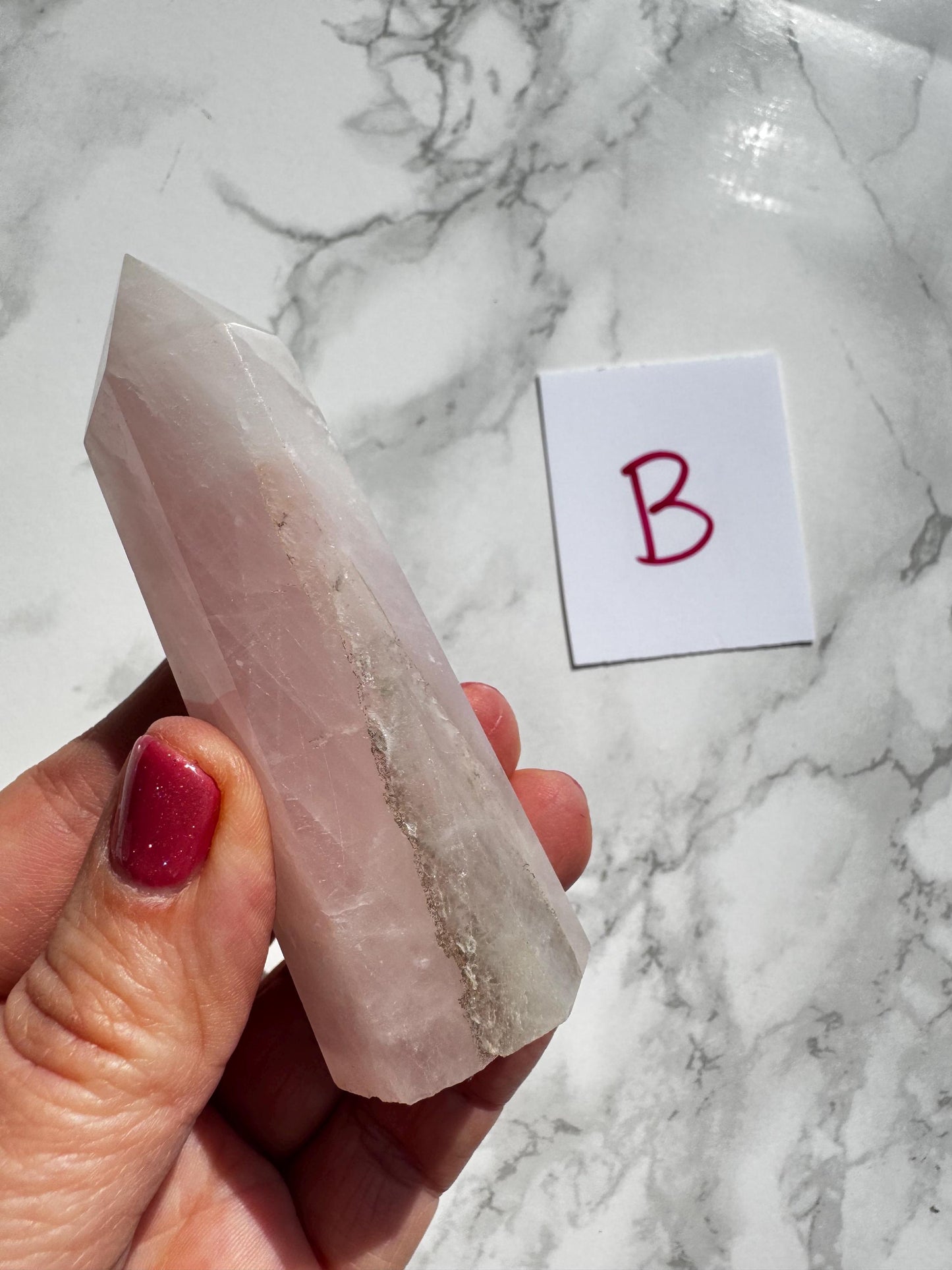 A hand holds a polished Rose Quartz Tower (3"–3.6")—YOU CHOOSE: Small A, B, C, or D—next to a white card with a red "B" on a marble surface.