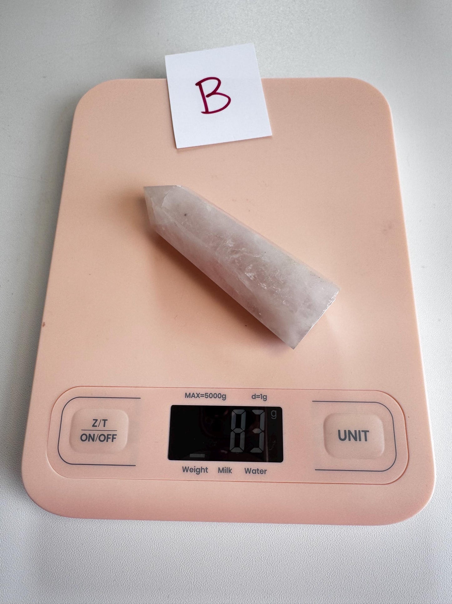 A pink digital kitchen scale reads 81 grams, holding a pointed white healing crystal and a card marked "B," often paired with Rose Quartz Towers (3"–3.6"), - YOU CHOOSE - Small A B C D.