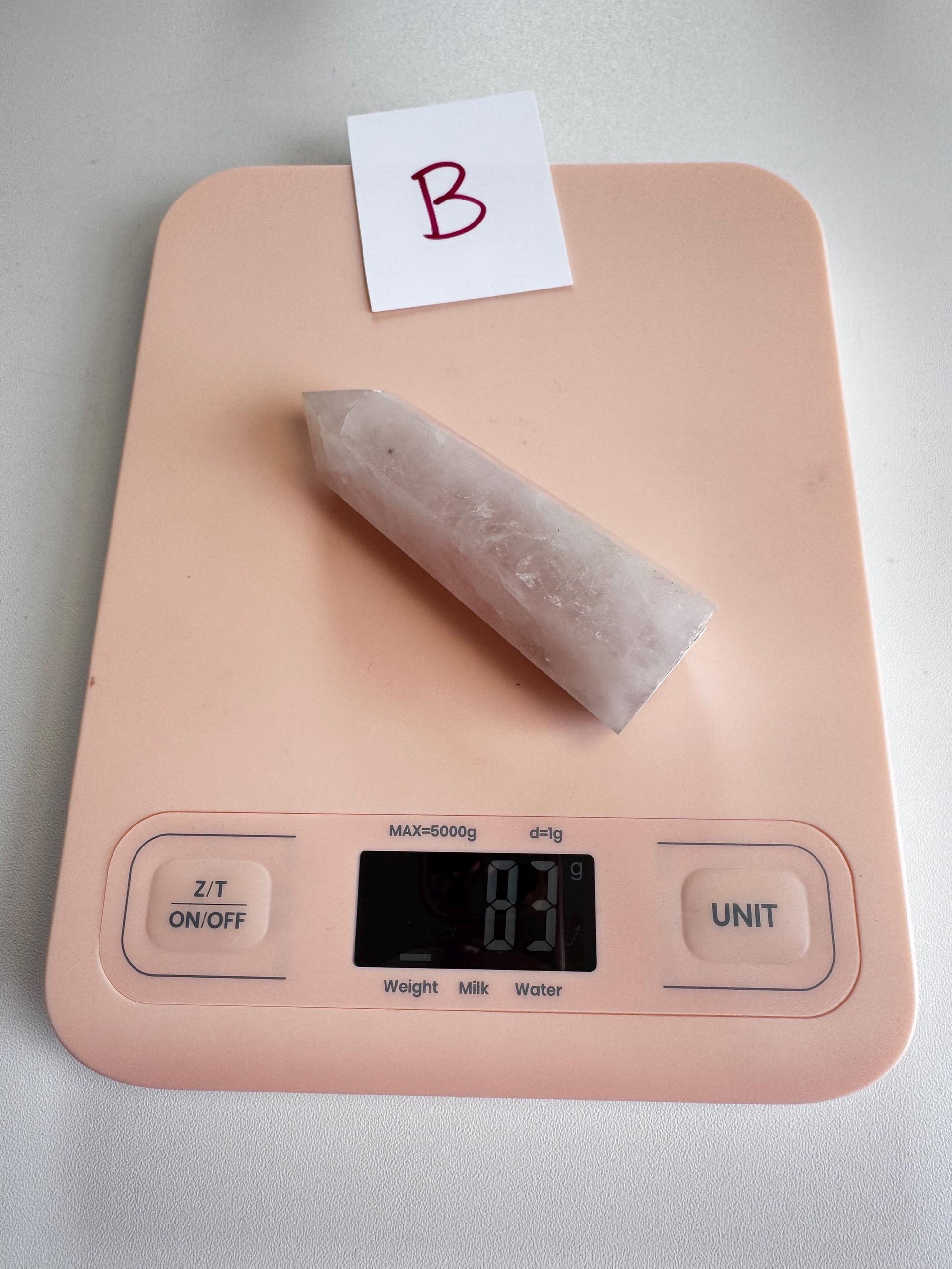 A pink digital kitchen scale reads 81 grams, holding a pointed white healing crystal and a card marked "B," often paired with Rose Quartz Towers (3"–3.6"), - YOU CHOOSE - Small A B C D.