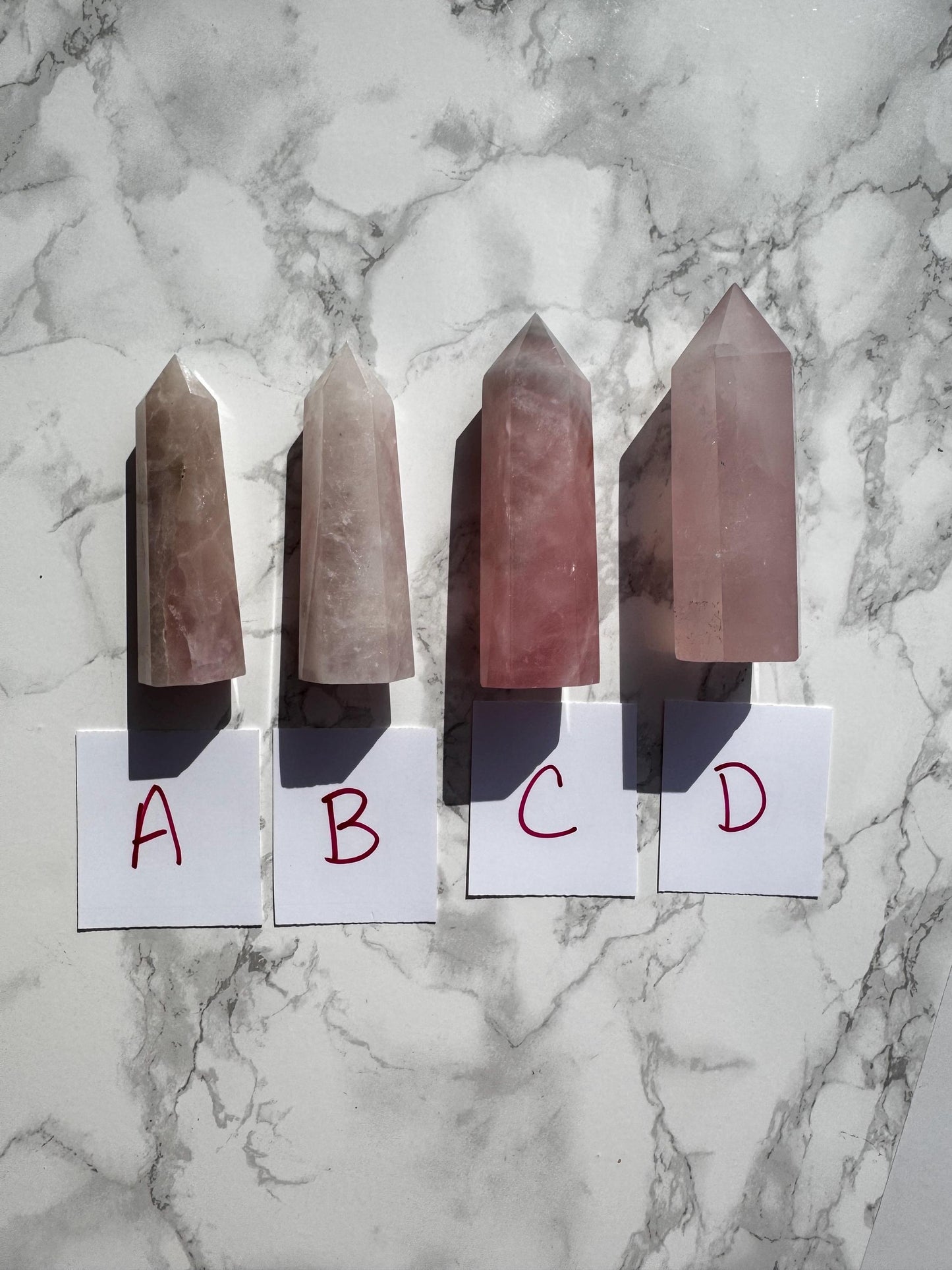Four Rose Quartz Towers (3"–3.6") labeled A, B, C, or D stand upright on marble with white cards—choose your perfect stone of unconditional love and healing.