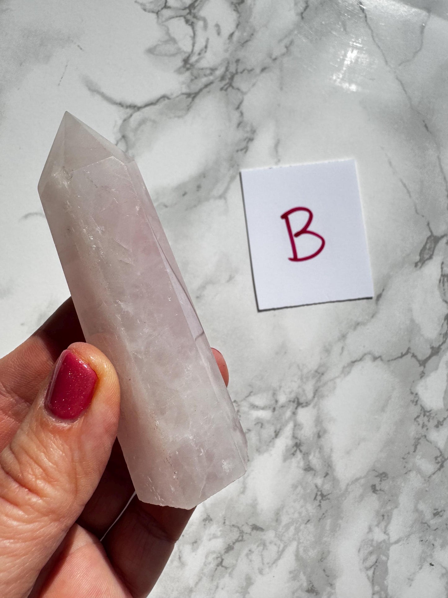 A hand with pink nail polish holds a 3–3.6" Rose Quartz Tower (“B”)—known for unconditional love—from the ‘YOU CHOOSE’ Small A B C D selection, next to a white “B” card on a marble surface.