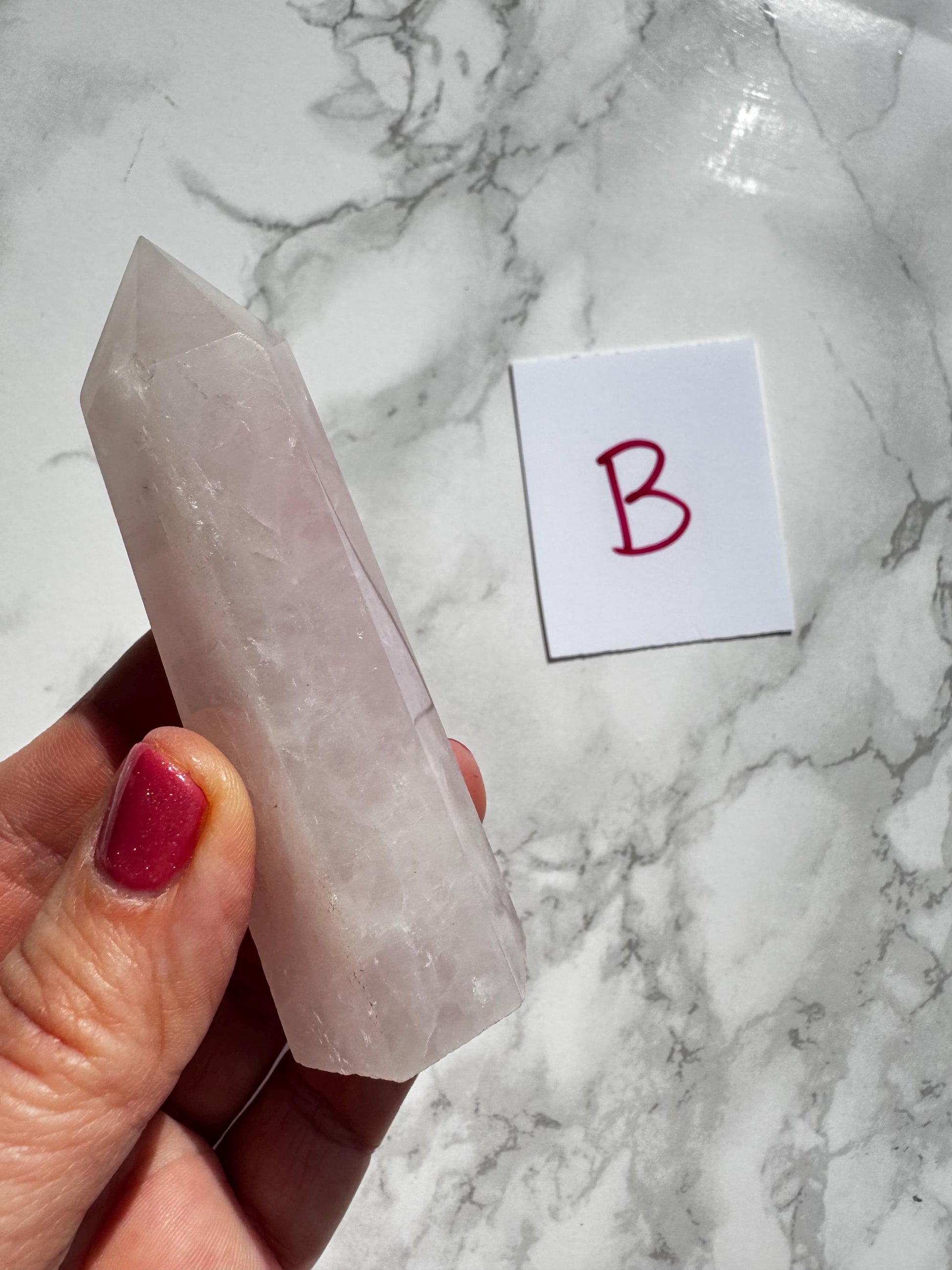 A hand with pink nail polish holds a 3–3.6" Rose Quartz Tower (“B”)—known for unconditional love—from the ‘YOU CHOOSE’ Small A B C D selection, next to a white “B” card on a marble surface.