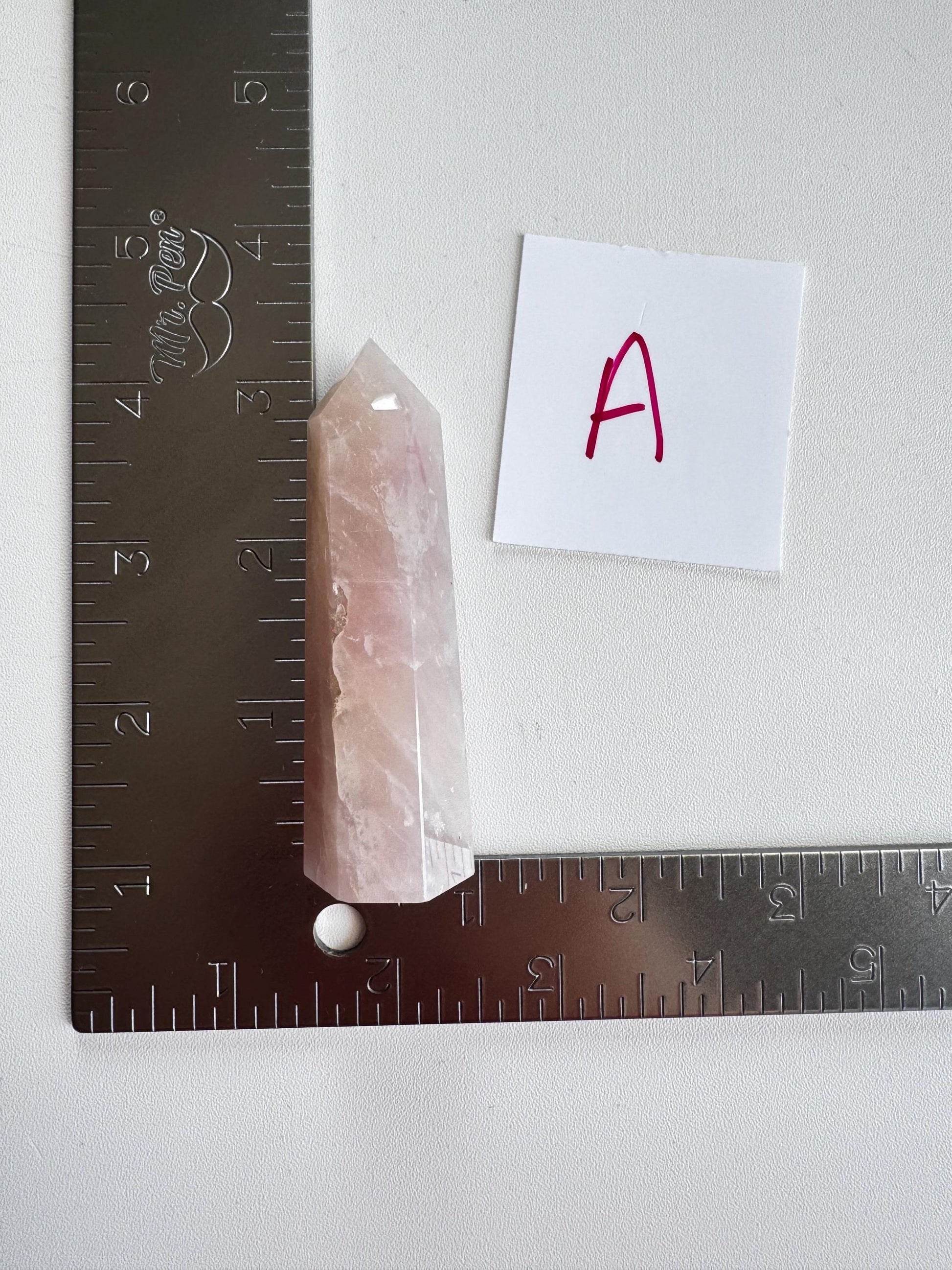 A Small 'A' Rose Quartz Tower (3"–3.6"), known for promoting unconditional love, is set at the corner of two L-shaped metal rulers, with a paper labeled "A" nearby.