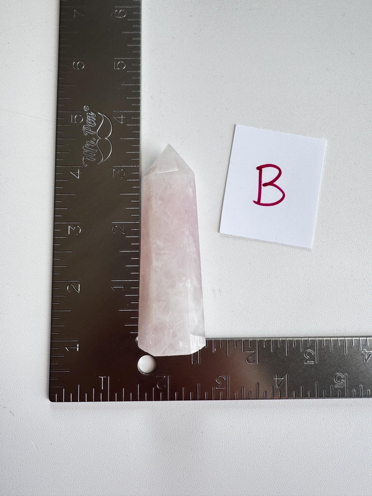 A Rose Quartz Tower (3"–3.6"), chosen from Small A, B, C, or D options, is positioned at the intersection of two metal rulers arranged in a right angle with a card labeled "B" nearby.