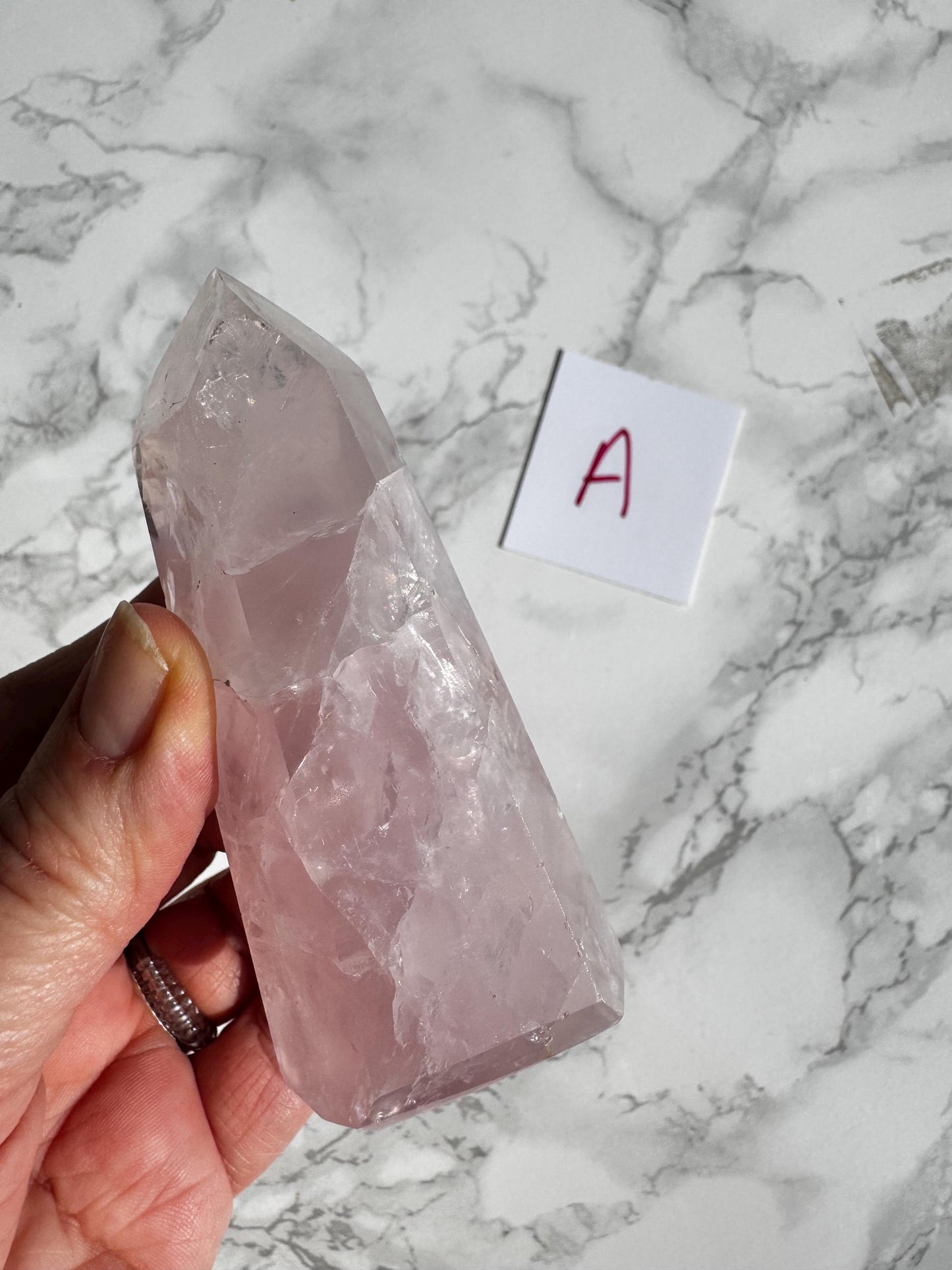 A hand holds a Nice Quality Rose Quartz Obelisk Tower from Brazil (3.6"-3.8") on marble, with a small “A” label nearby—an elegant display for healing crystal lovers. YOU CHOOSE your unique obelisk!.