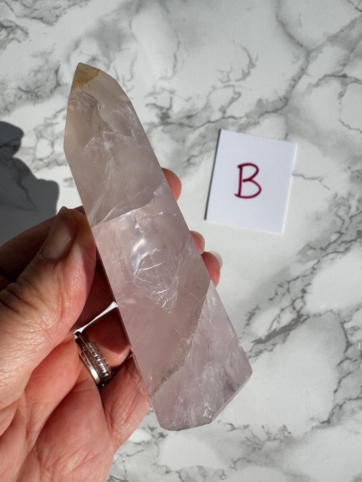 A hand holds a Nice Quality Rose Quartz Obelisk Tower from Brazil (3.6" - 3.8") with a white "B" card on a marble background.