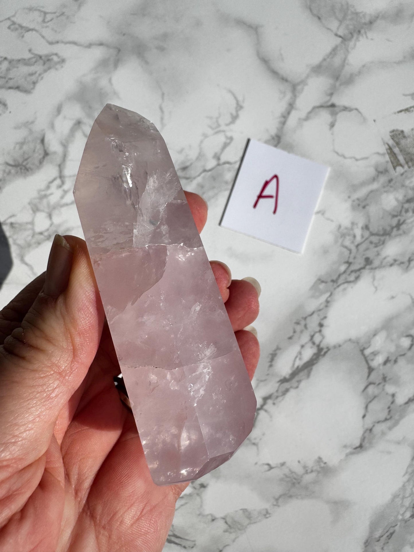 A hand holds a Nice Quality Rose Quartz Obelisk Tower from Brazil (3.6" - 3.8"); a small white paper labeled “A” in red ink rests on the marble surface nearby.