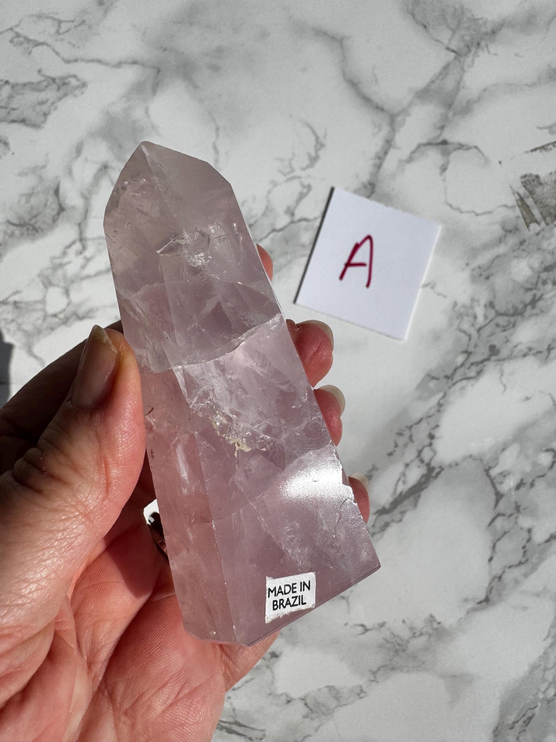 A hand displays a polished Rose Quartz Obelisk Tower (3.6"–3.8") labeled "Nice Quality, Made in Brazil"; a white card with a red "A" rests on marble, highlighting the allure of these healing crystals—YOU CHOOSE your favorite.