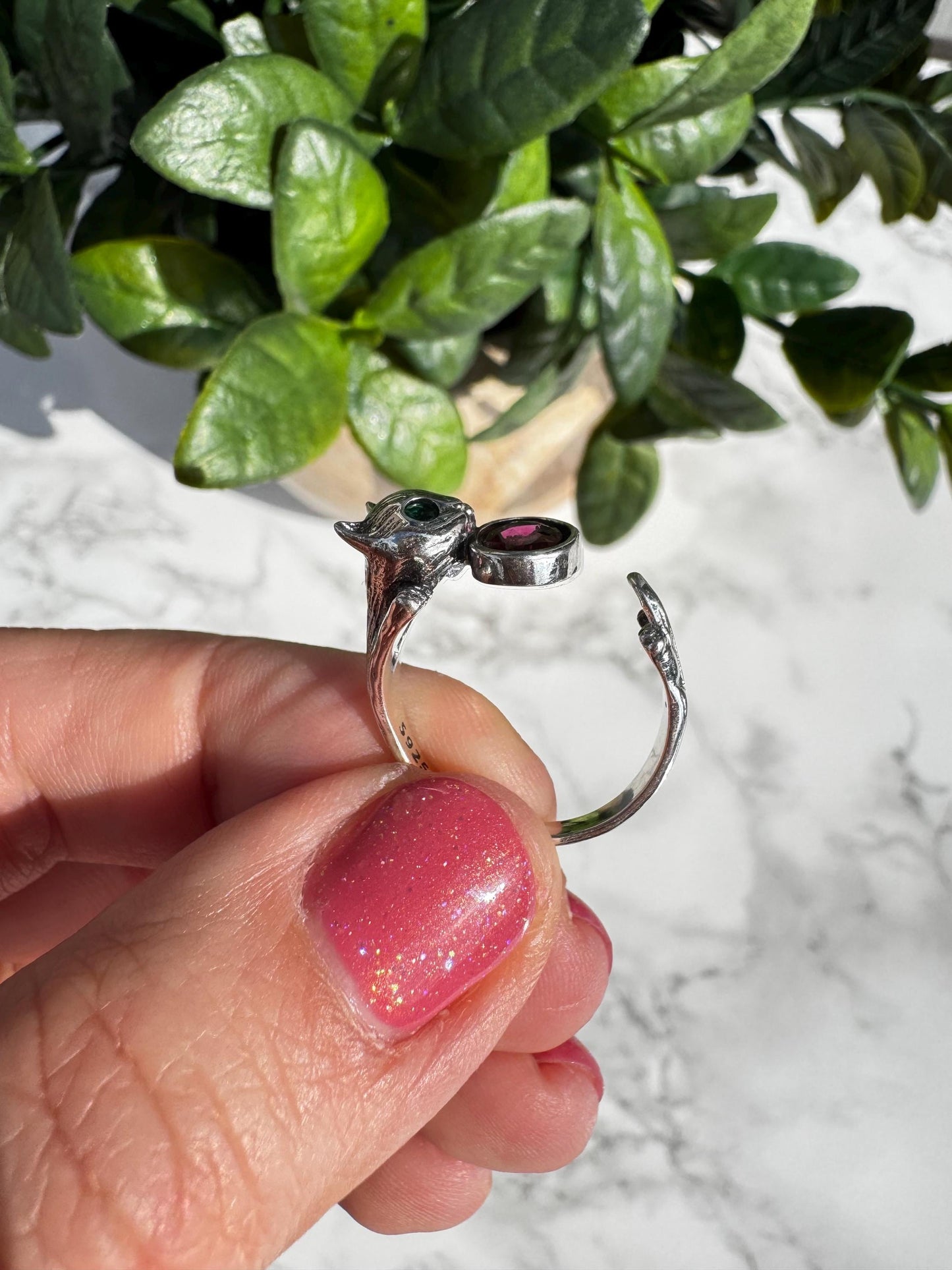 A hand with pink glittery nail polish holds a Sterling Silver Cat Ring with Pink Tourmaline Gemstone (Size 6.5, adjustable), while a green potted plant sits in the background.