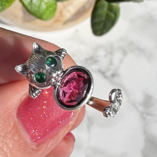 The Sterling Silver Cat Ring with Pink Tourmaline Gemstone (Size 6.5, Adjustable) features green gemstone eyes and an oval tourmaline body—an eye-catching, playful piece of jewelry perfect for cat lovers and crystal healing enthusiasts.