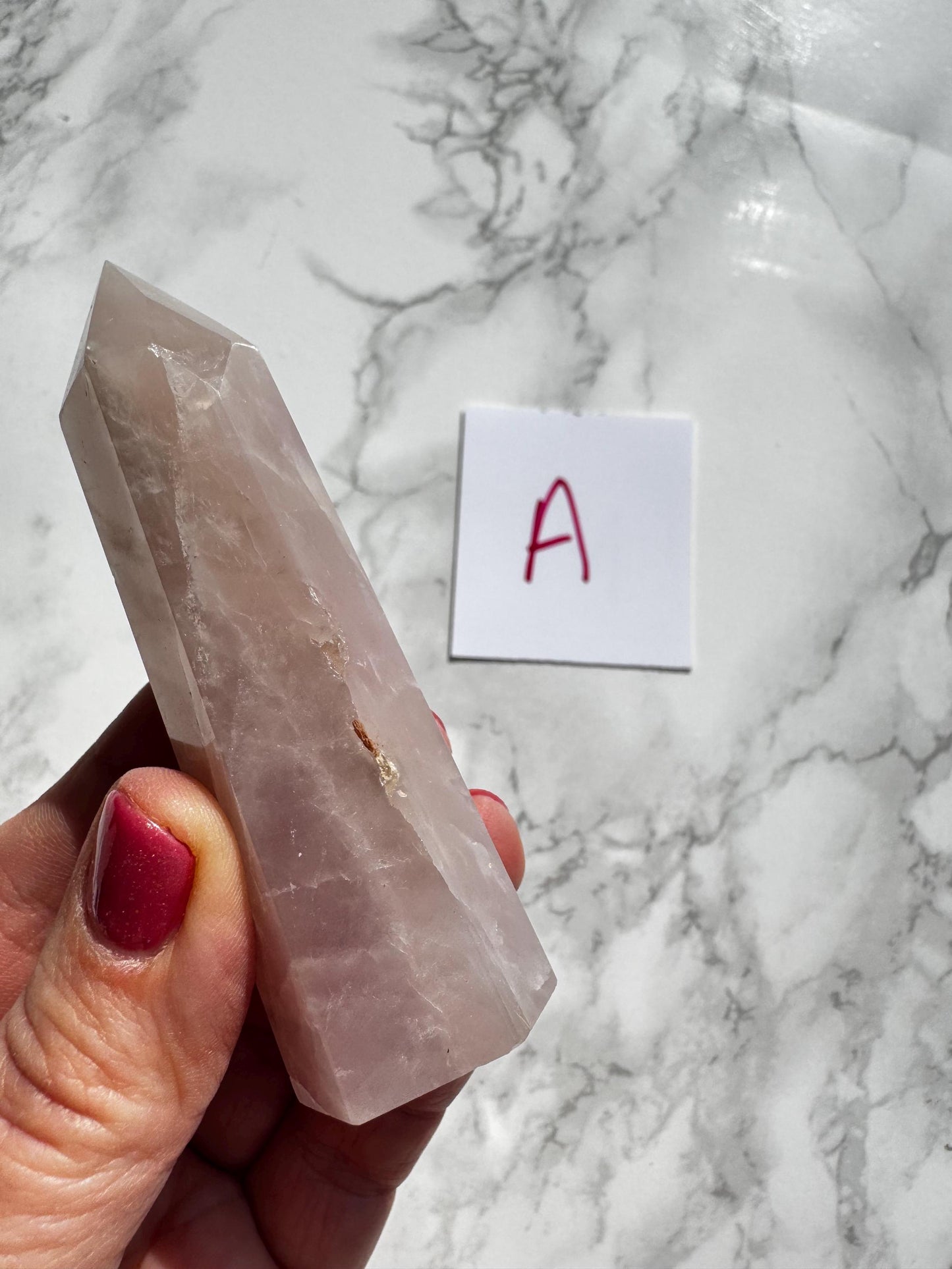 A hand holds a pointed, translucent Rose Quartz Tower (3"–3.6"). In the background, a white card marked "A" rests on marble—one of several YOU CHOOSE Rose Quartz Towers (Small A B C D).