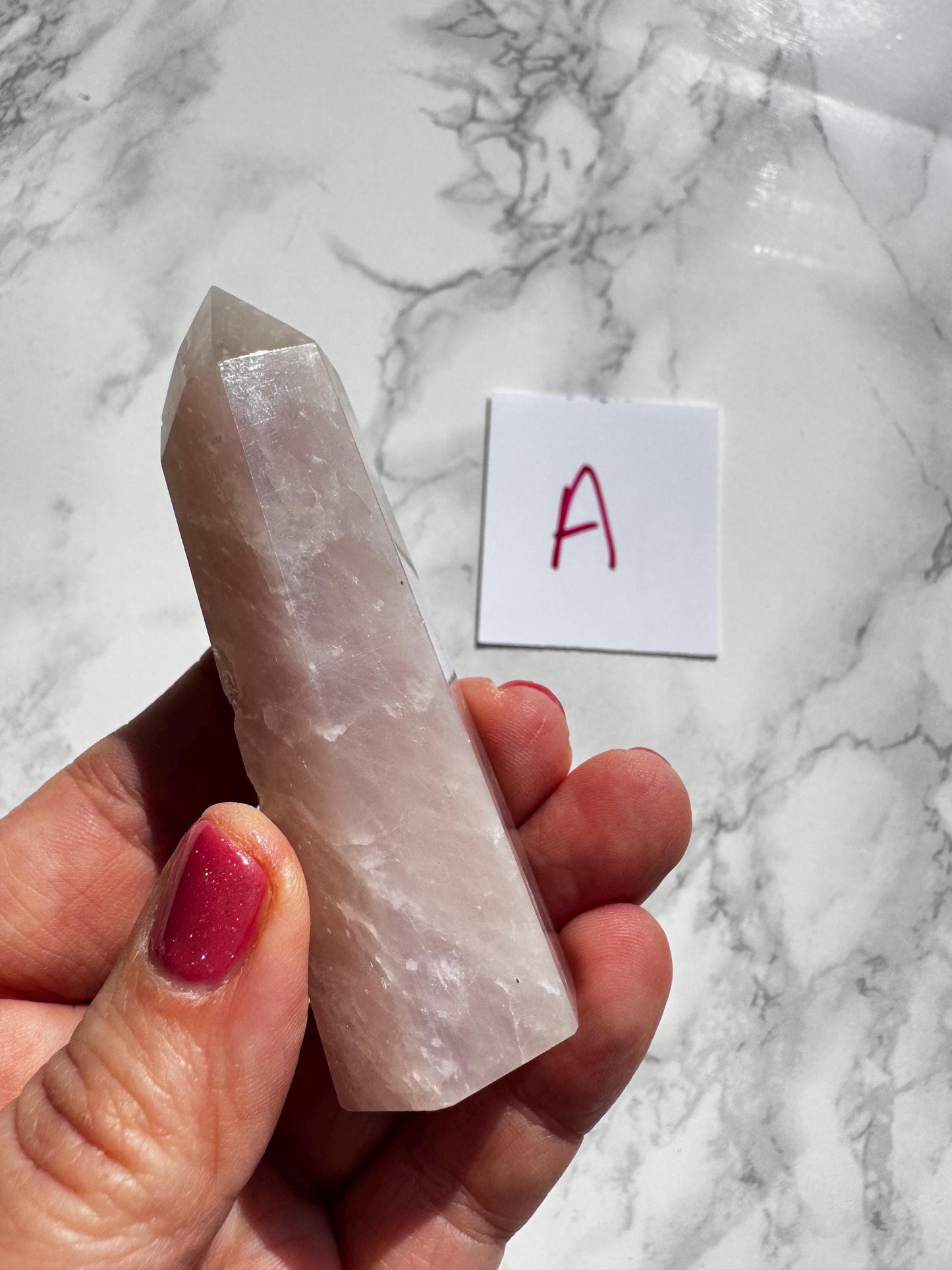 A hand holds a 3"-3.6" Rose Quartz Tower (Small A) from the "YOU CHOOSE - Small A B C D" selection, next to a white card labeled "A" in red marker on a marble surface.