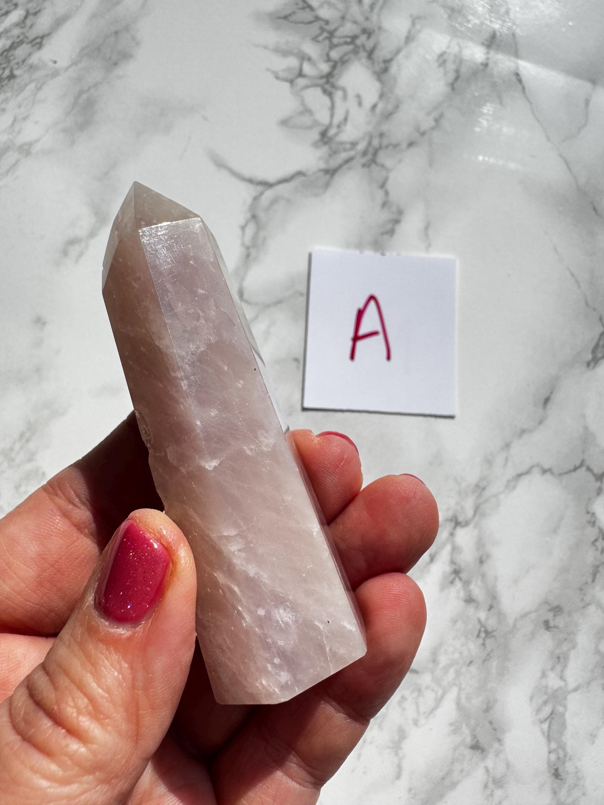 A hand holds a 3"-3.6" Rose Quartz Tower (Small A) from the "YOU CHOOSE - Small A B C D" selection, next to a white card labeled "A" in red marker on a marble surface.